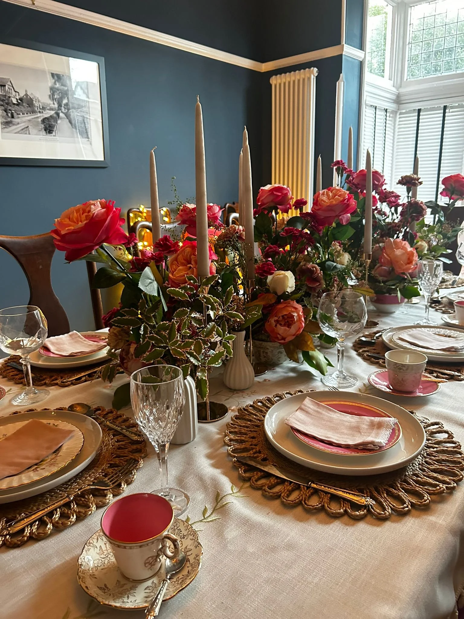 A dining table set for a meal with floral centerpiece, candles, fine china, crystal glasses, and place settings on woven place mats, in a room with blue walls and large windows.