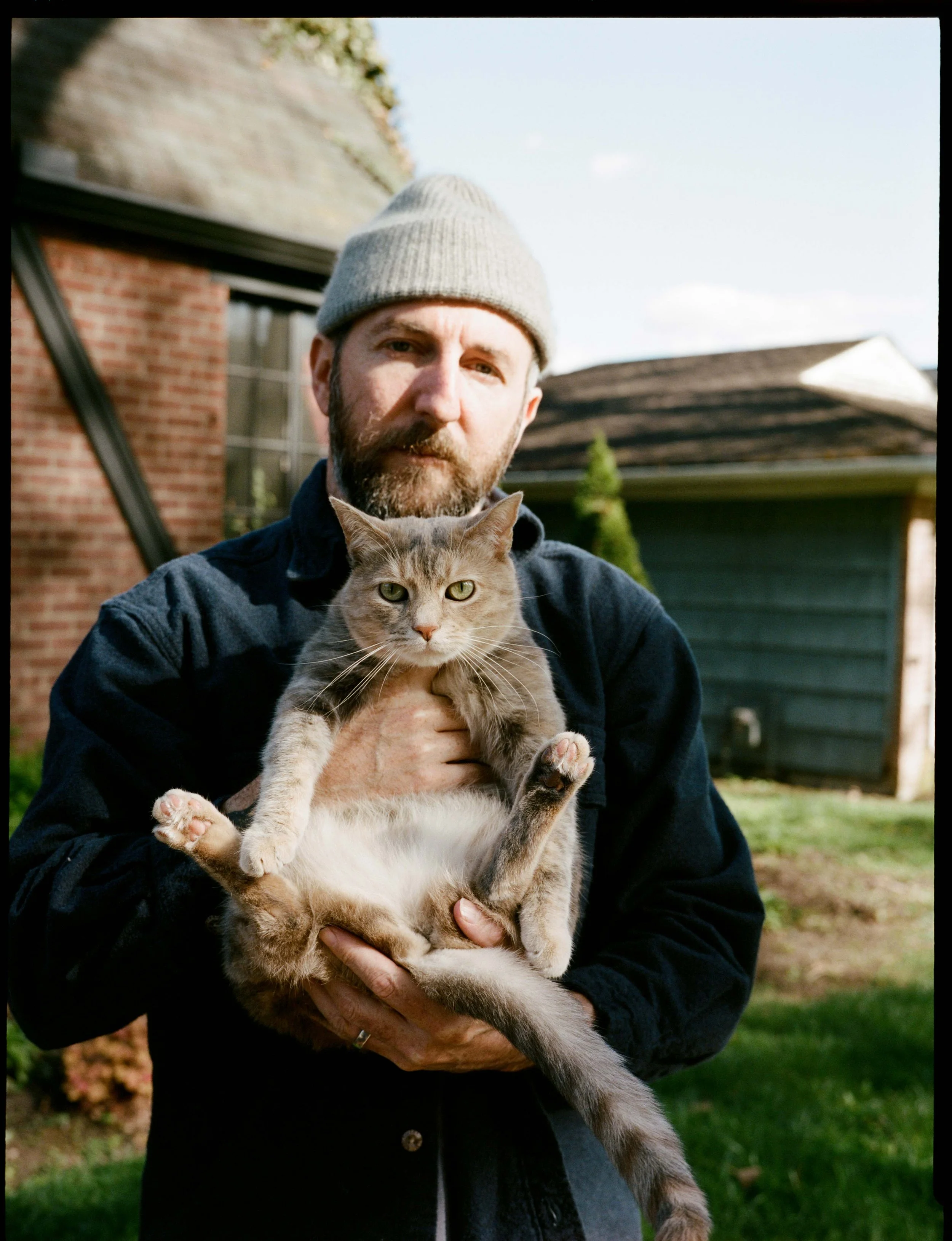 A man with a beard and gray beanie holding a tabby cat outside in a backyard with a brick building and a wooden shed in the background.