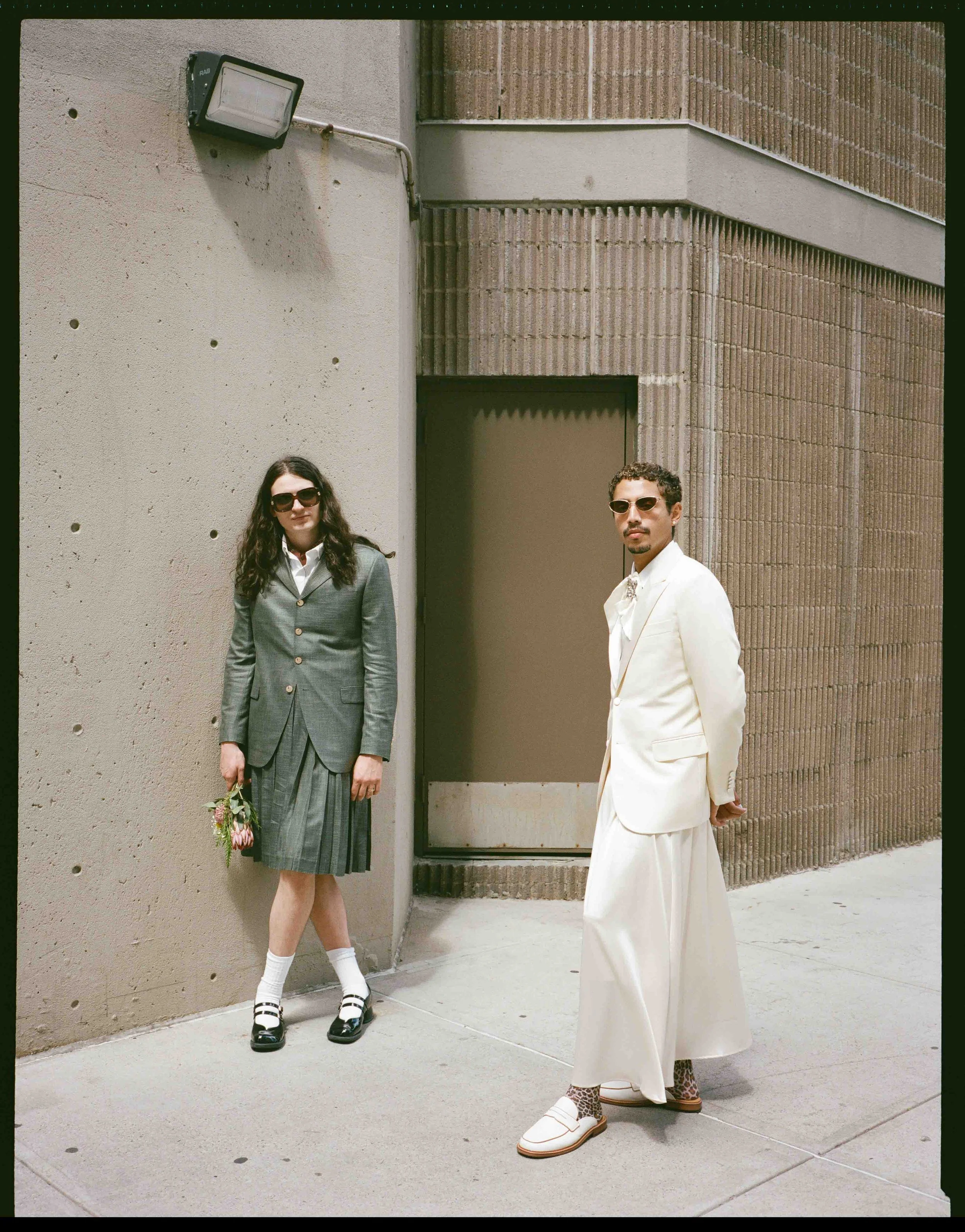 Two people standing outside a building, dressed in vintage-style clothing, with a concrete wall and brick building in the background.