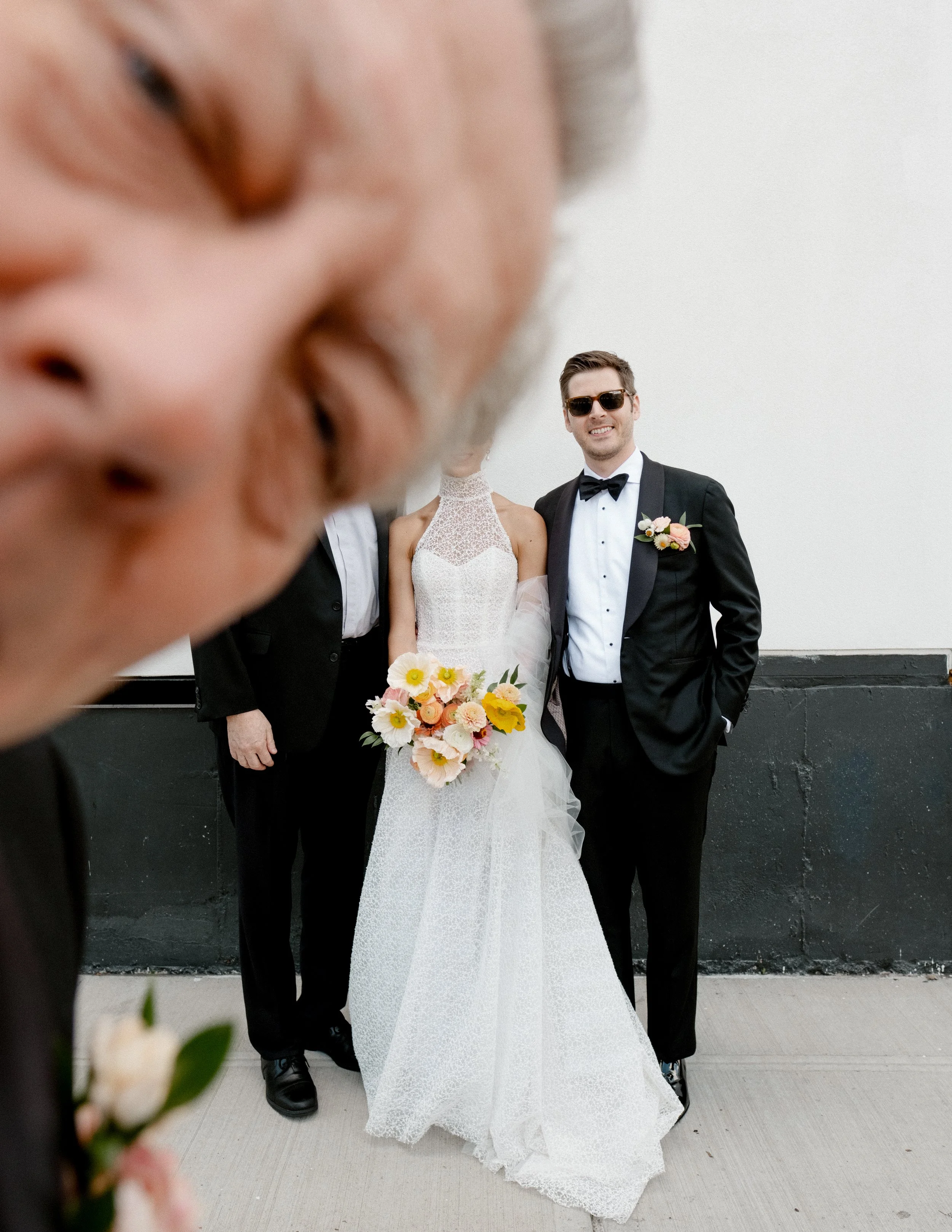 Photo of a wedding scene with a bride holding a bouquet of flowers and three men in formal attire, one wearing sunglasses, standing against a plain white wall. In the foreground, a person's face is close to the camera.