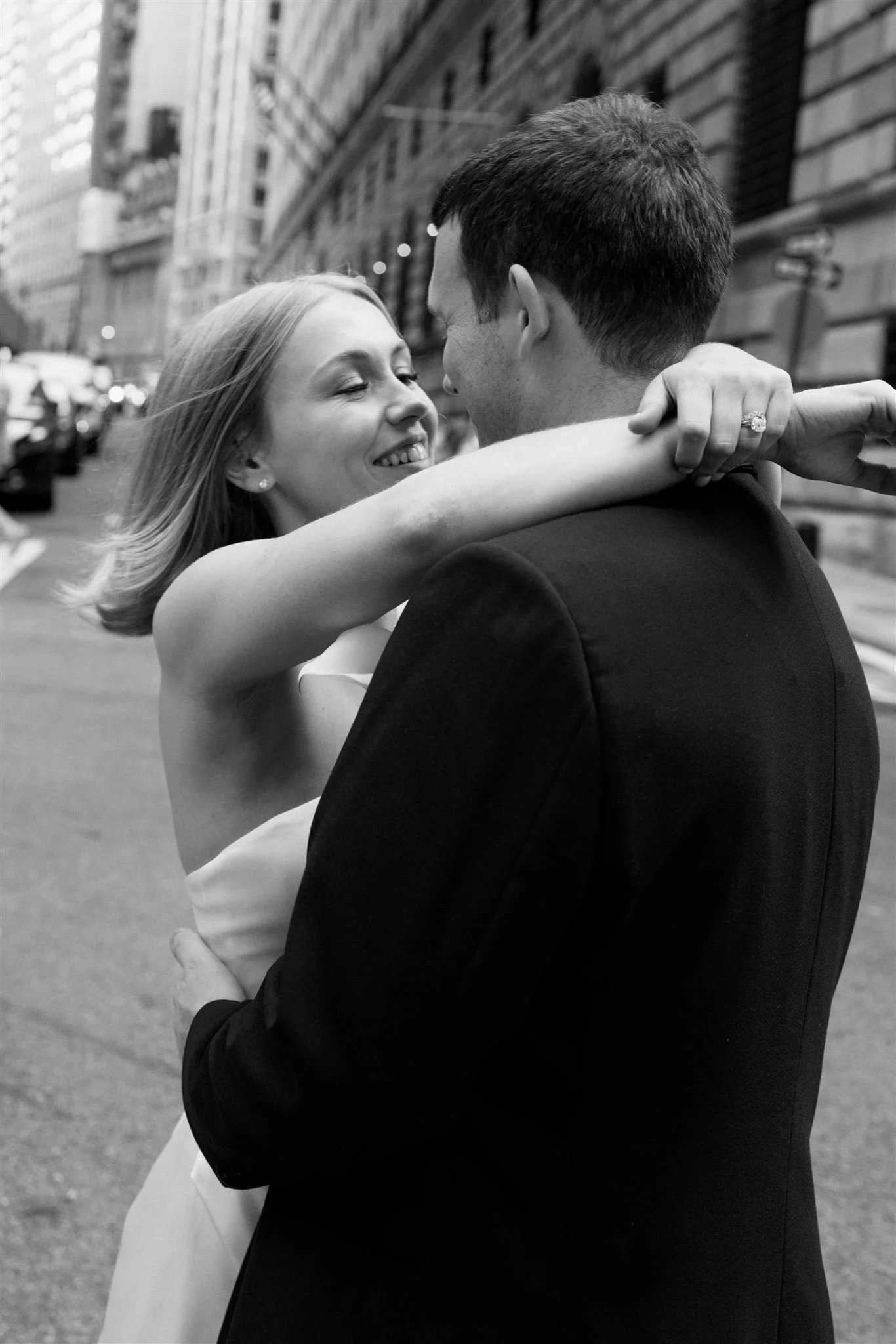 A couple dancing closely on a city street, smiling and looking into each other's eyes, with buildings and parked cars in the background.