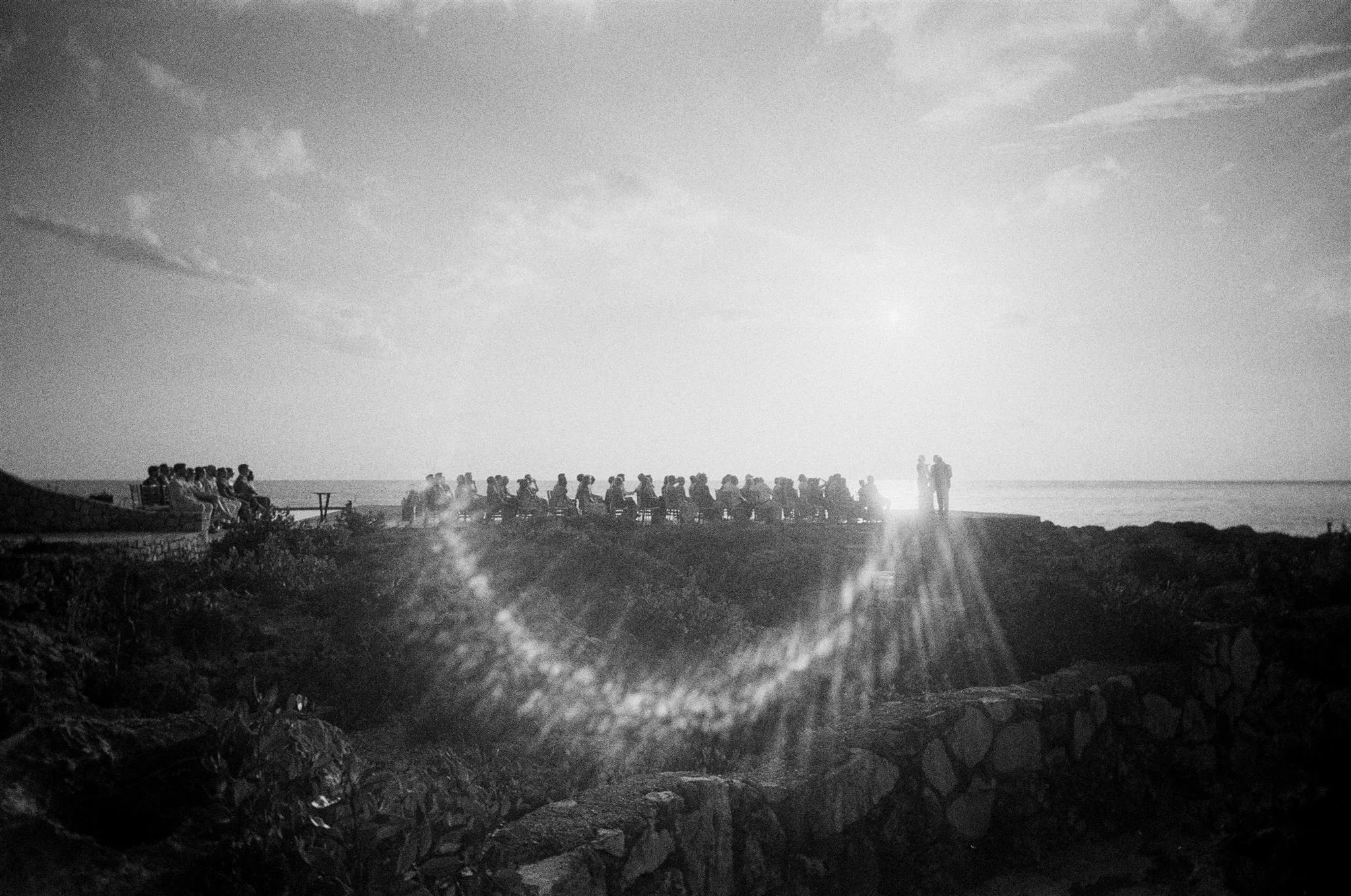 Black and white photograph of people sitting and standing along a coastal overlook, with the ocean and sky in the background, and the sun low on the horizon.