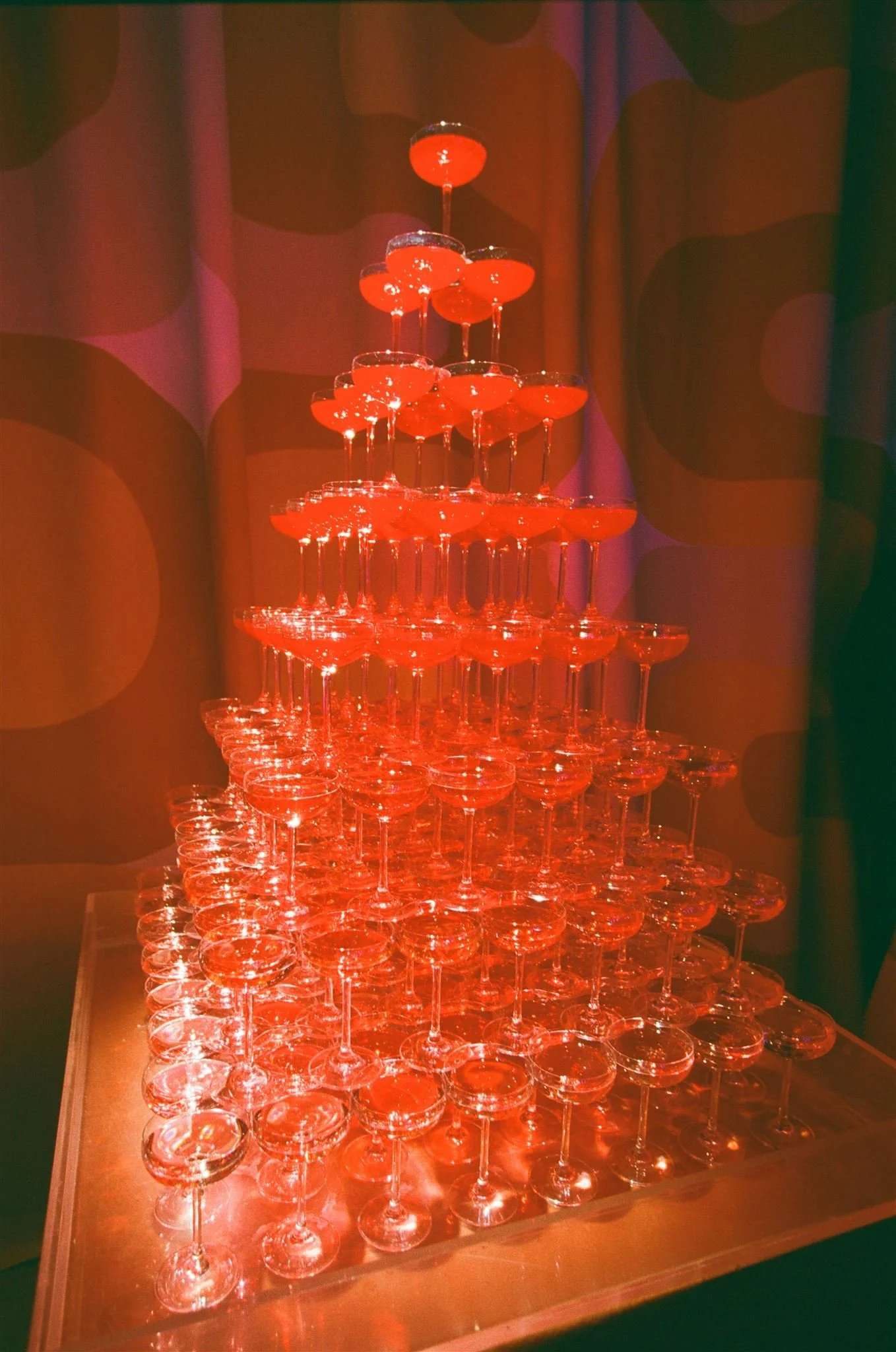 A pyramid of stacked champagne glasses with red drinks, set against a colorful, abstract background.