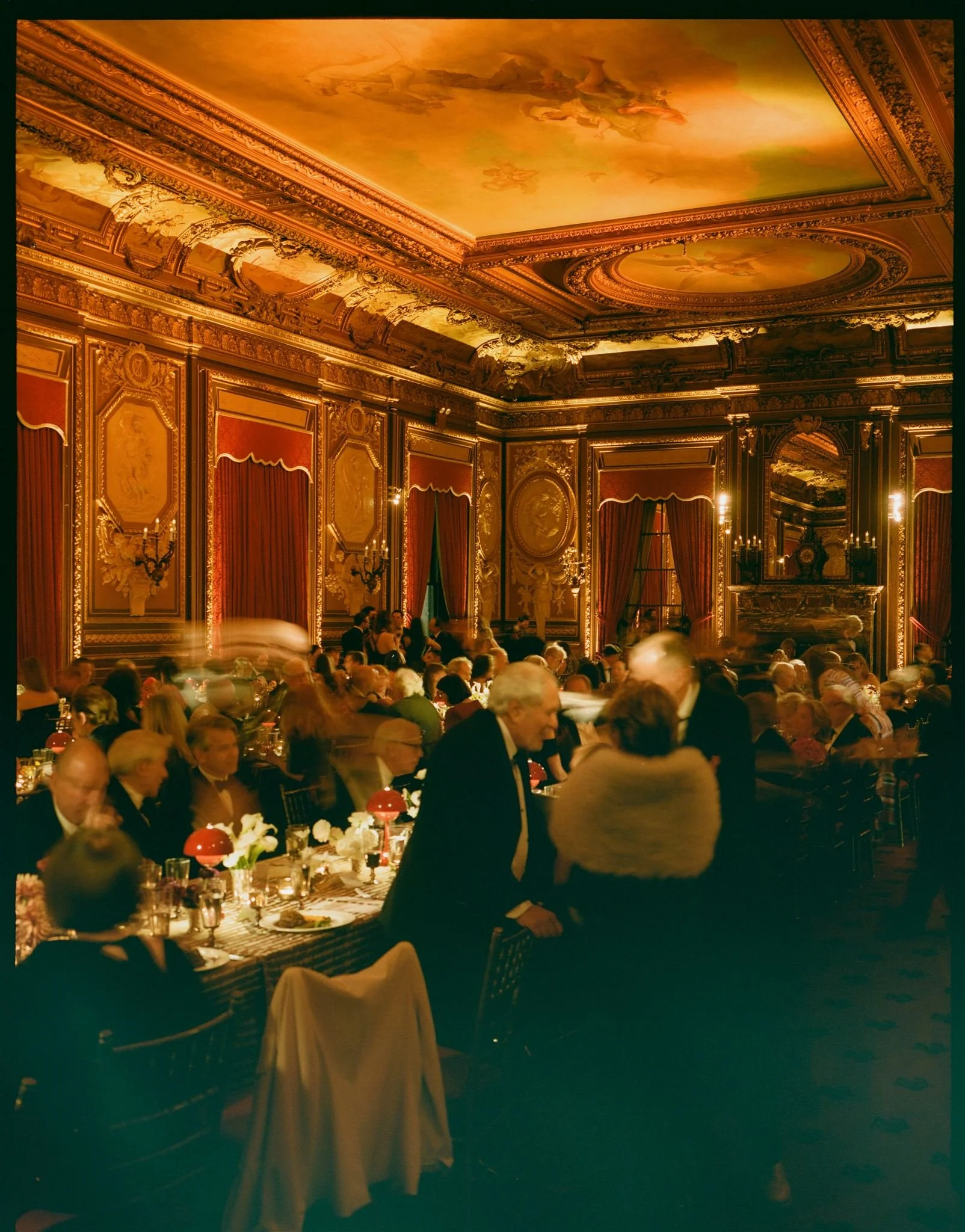 An elegant banquet hall with ornate gold and red decor, high ceilings with painted murals, and a large mirror over a fireplace. People are seated at long tables, engaged in conversation, with soft lighting creating a warm atmosphere.