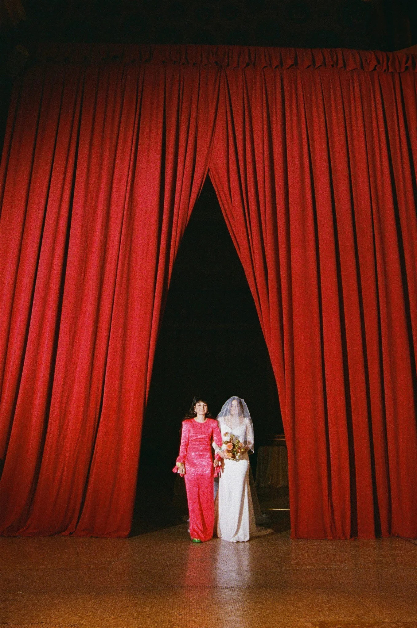 Two women, one in a pink dress and the other in a white wedding gown with a veil, holding a bouquet, walking out from behind red theater curtains.