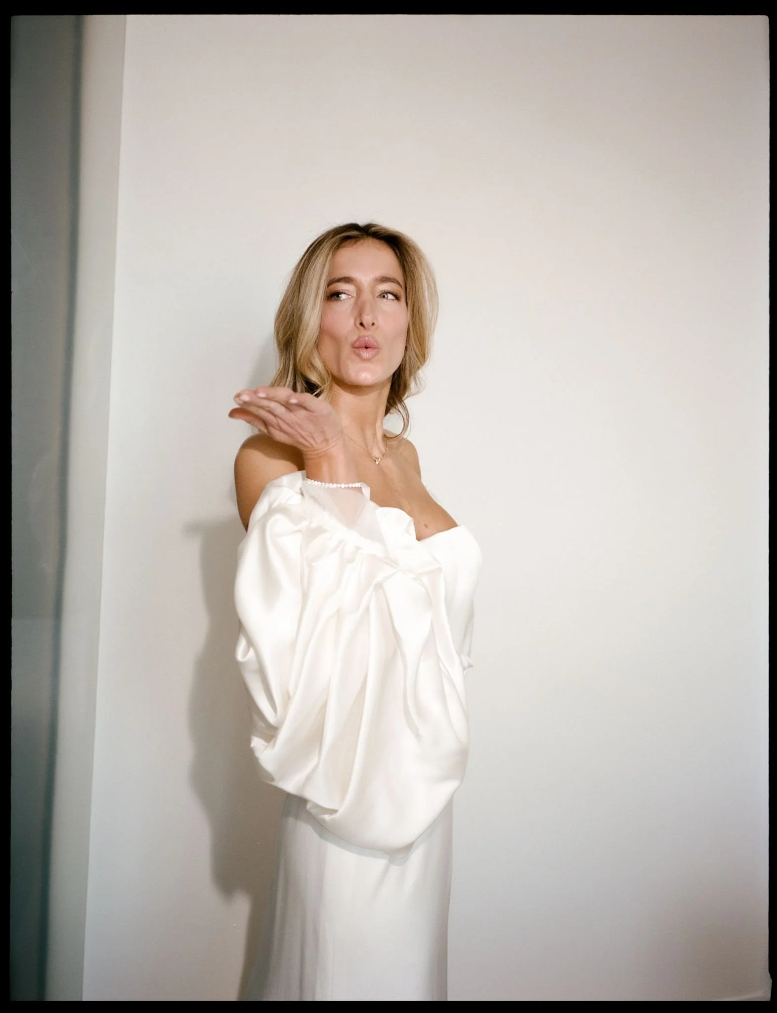 A Brooklyn bride with blonde hair in an off-the-shoulder white dress blowing a kiss in front of a plain white wall.