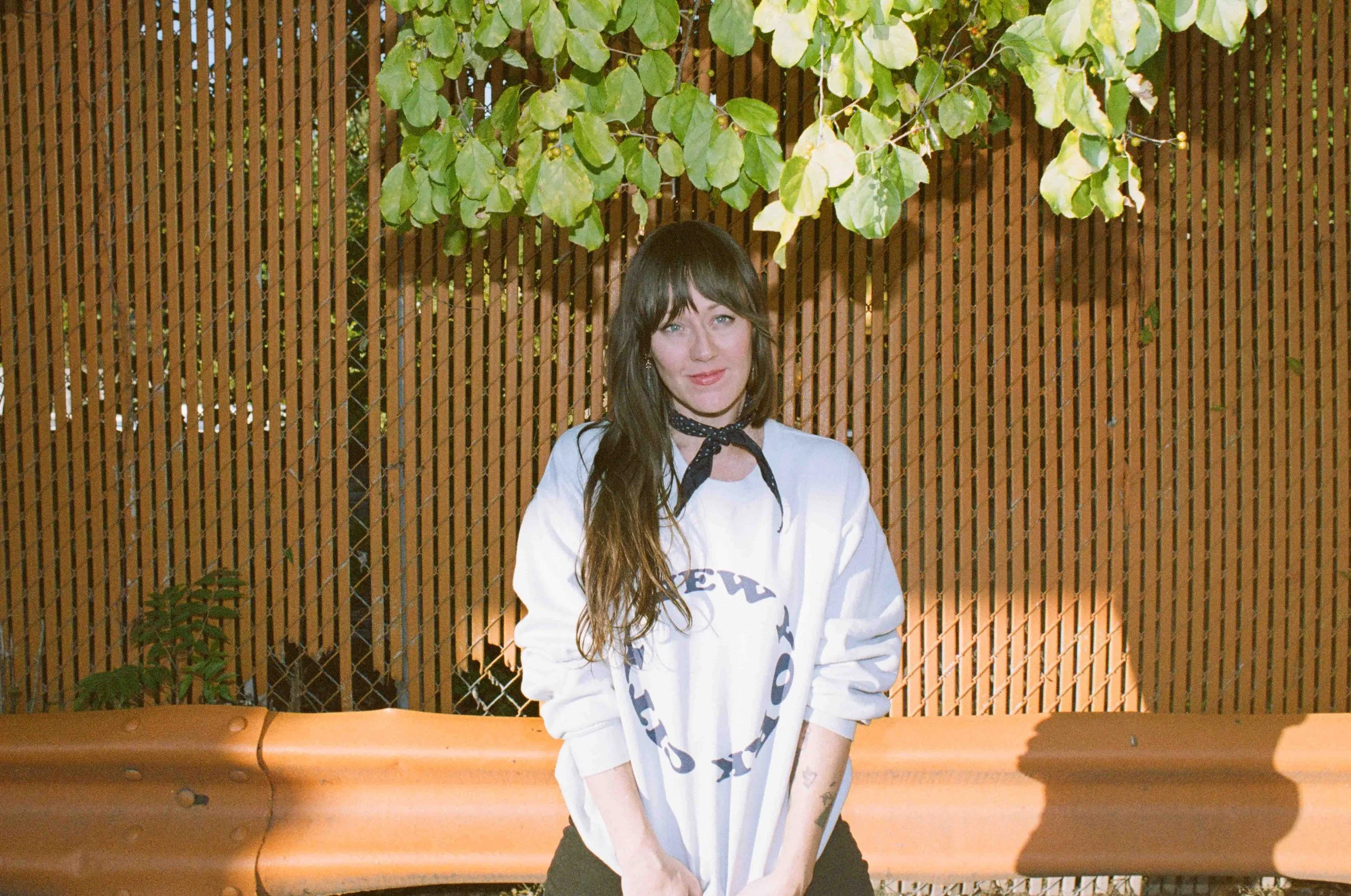 A young woman with long brown hair, wearing a white shirt and a black bandana, sitting on a brown park bench in front of a wooden fence with green leafy branches overhead.