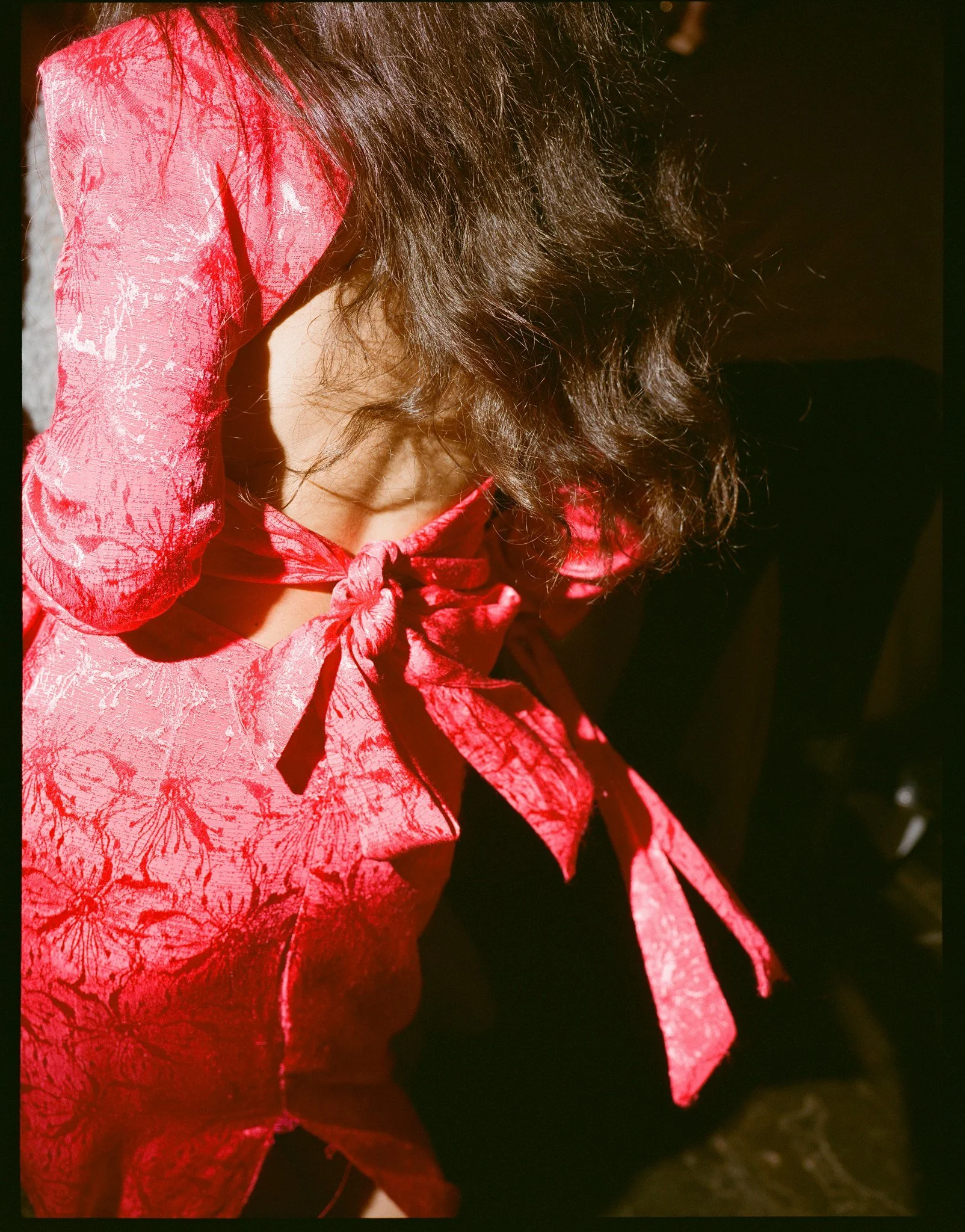 Close-up of a woman with dark, curly hair wearing a bright red dress with a bow at the collar, illuminated by sunlight.