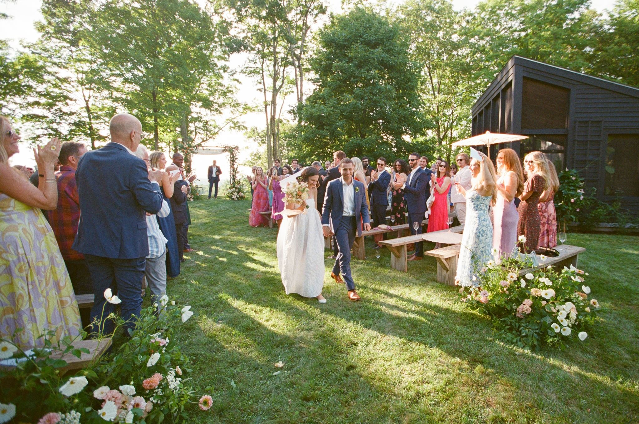 A wedding ceremony outdoors with guests clapping and smiling, surrounded by trees and flowers.