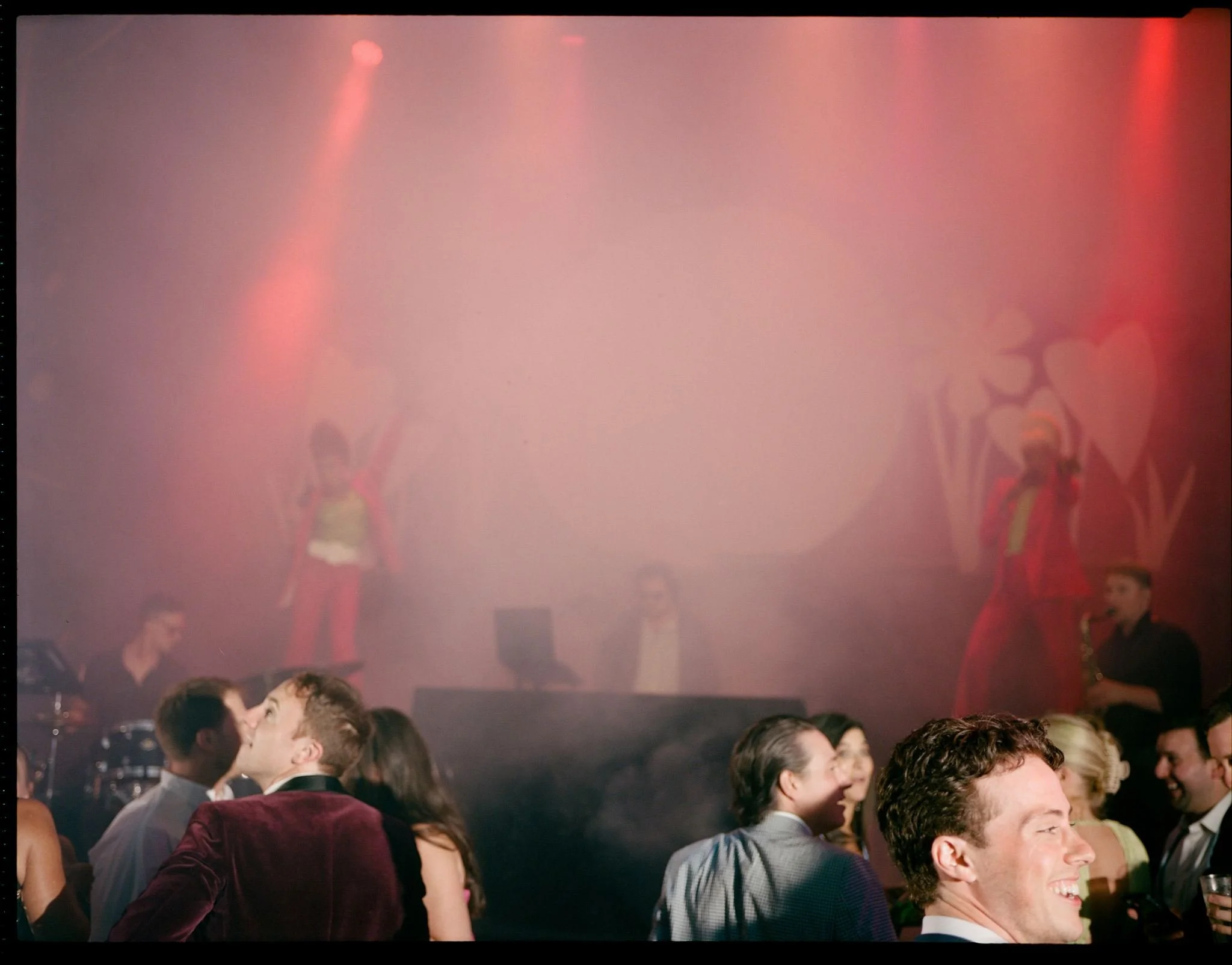 Guests dancing and socializing at a lively party with performers on stage, colorful lighting, and decorated backdrop.