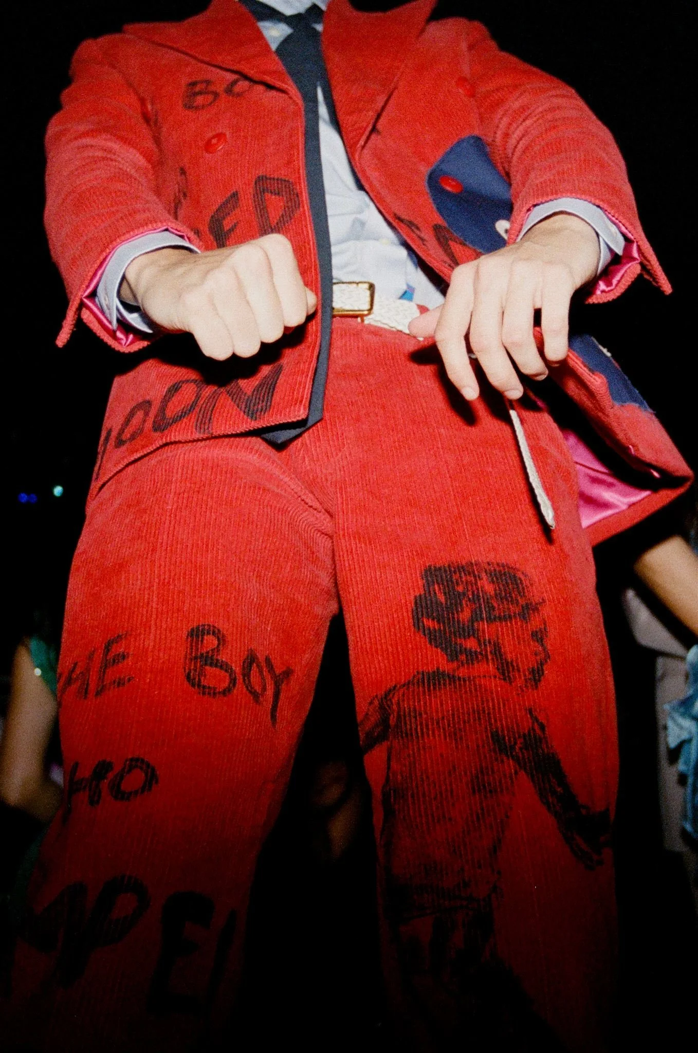 A person wearing a red jacket and red pants with black writing and drawings on them, standing with fists clenched.