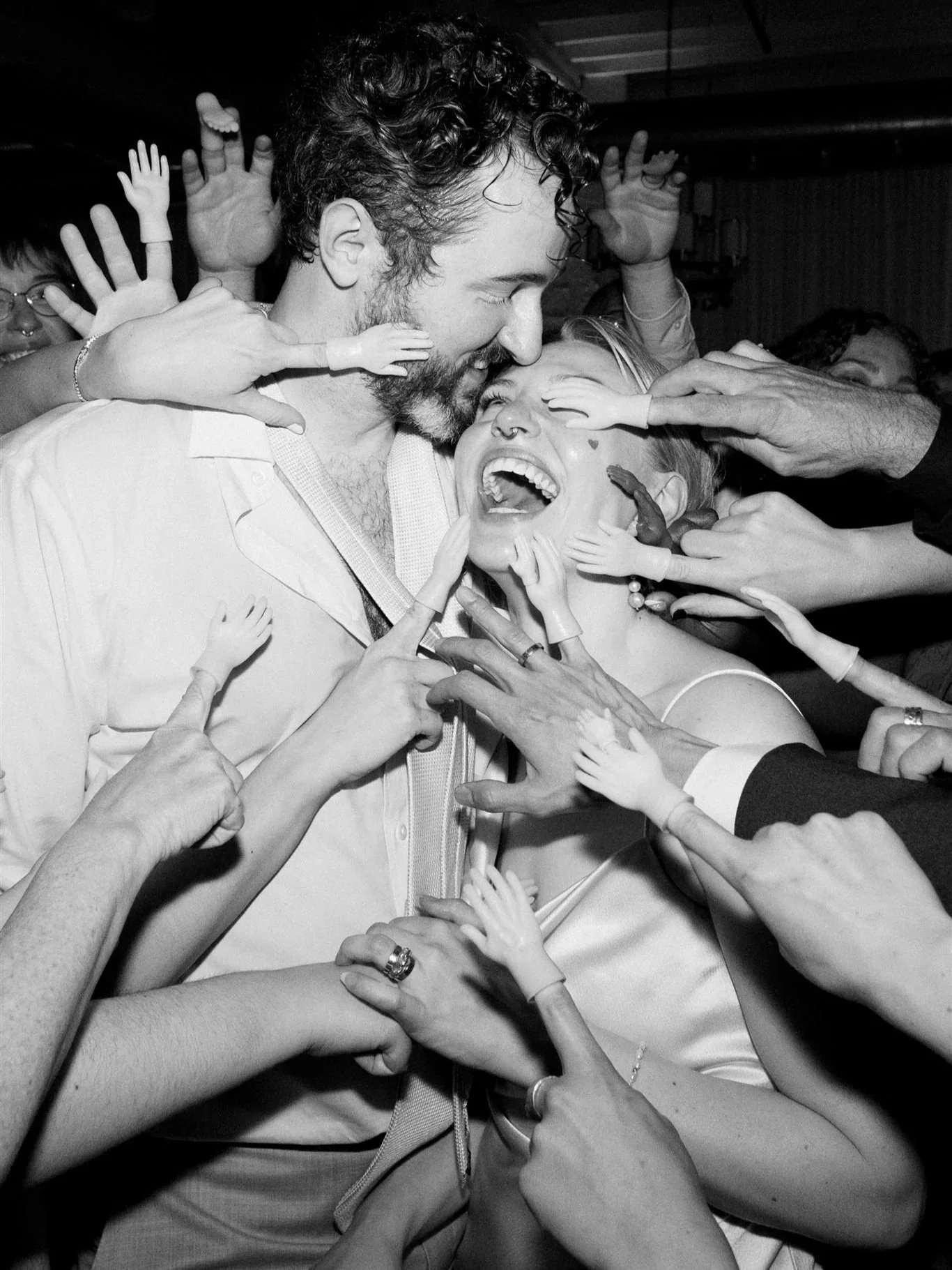 A black and white photo of a man and woman smiling and laughing, surrounded by multiple hands reaching towards them, some touching their faces, in a joyful embrace at what appears to be a lively celebration or party.