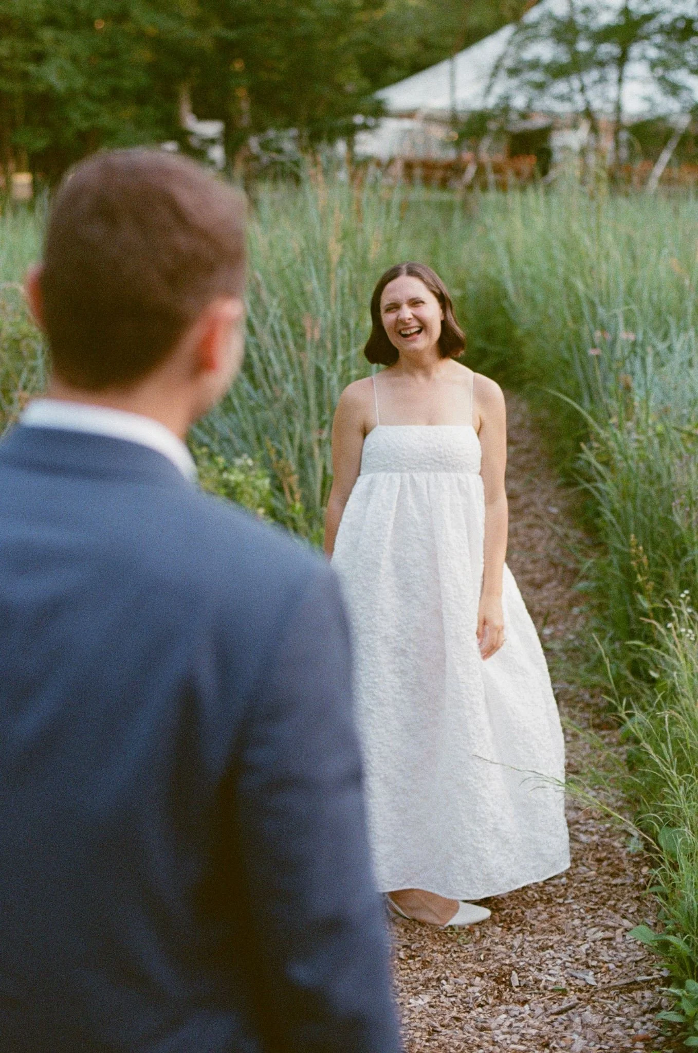Smiling woman in a white dress standing on a garden path, view of a man in a suit back to the camera in the foreground.