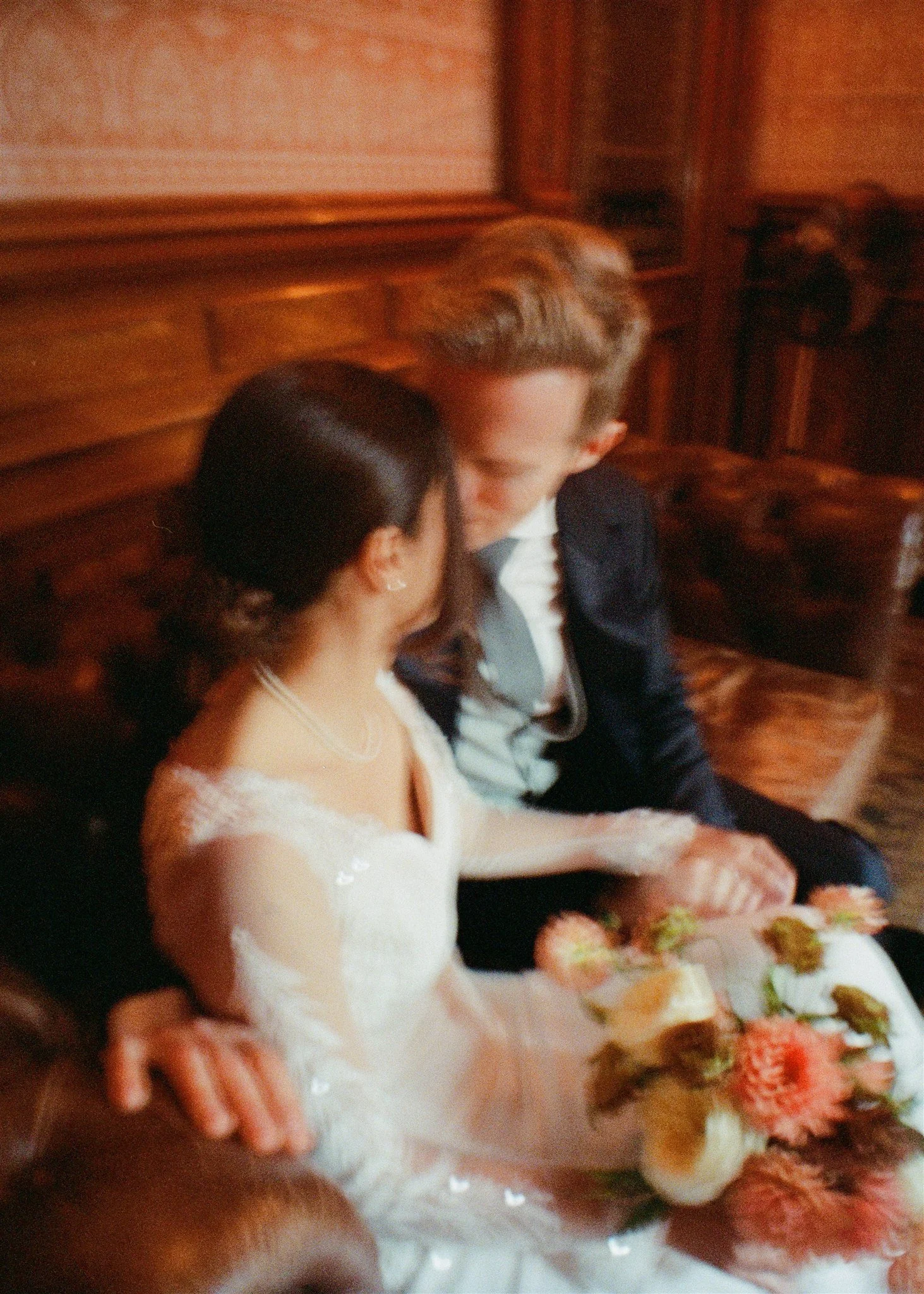 A blurry photo of a bride and groom sitting closely together, with the bride in a white wedding dress holding a bouquet, and the groom in a dark suit, in a wood-paneled room.