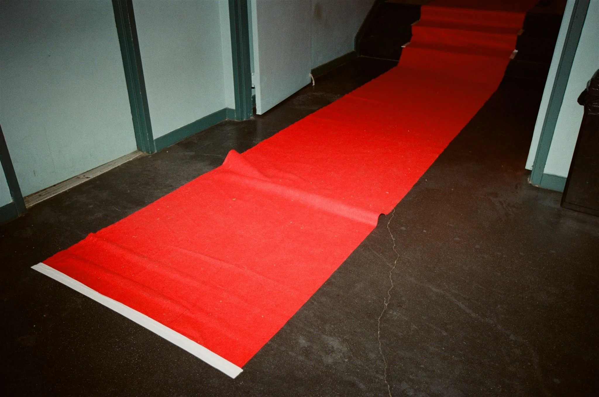 A red carpet runner laid out on a dark, cracked floor leading toward a staircase in a building interior.