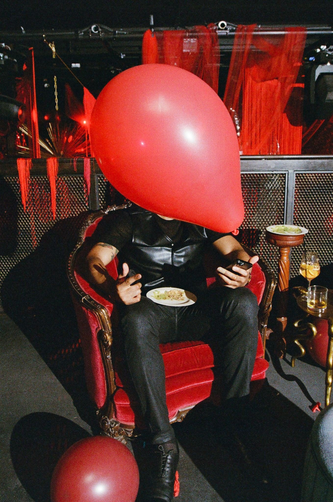 Person sitting on a red, ornate chair at a party, with a large red heart-shaped balloon covering their head, using a smartphone and holding a plate of food.