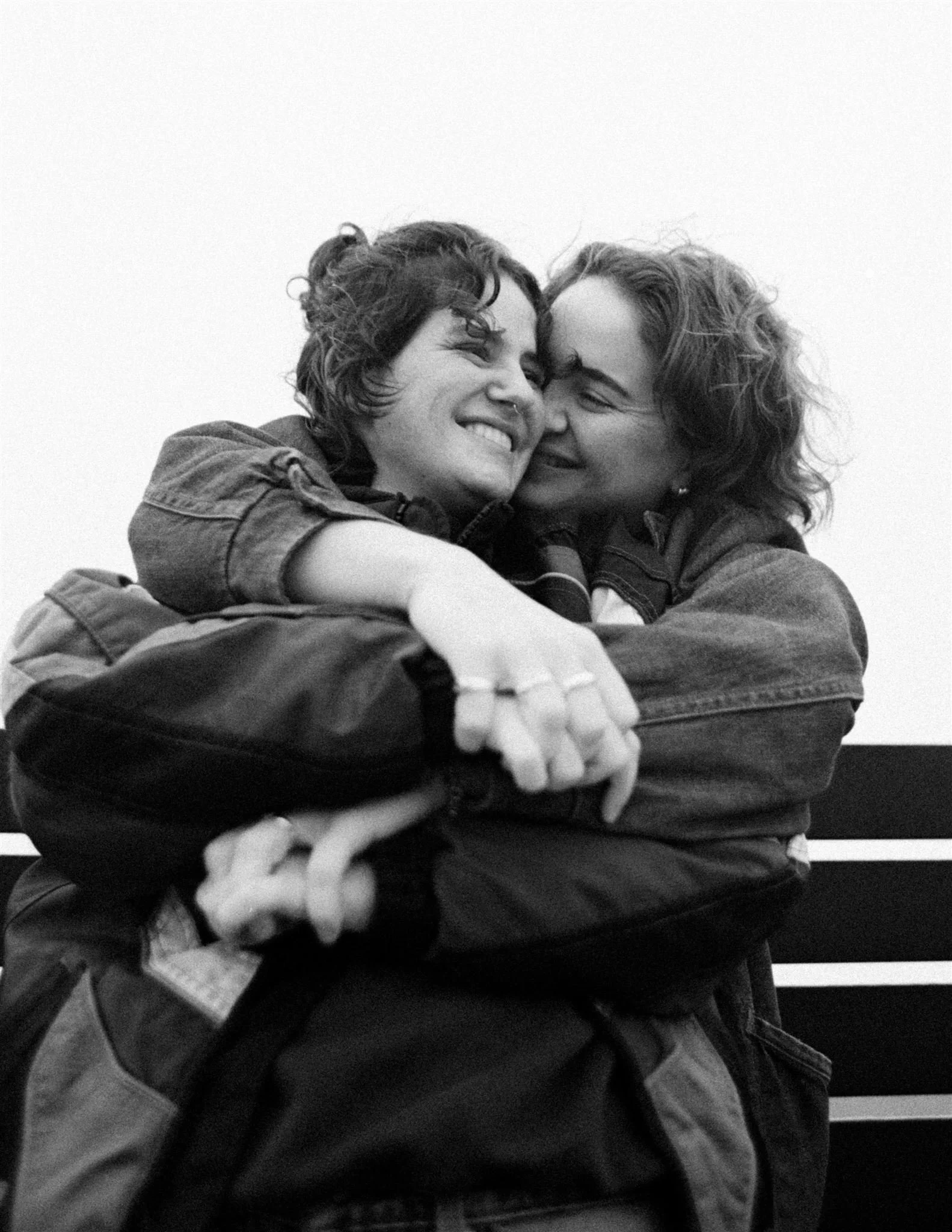 Two women embracing and smiling at each other, dressed in casual jackets, black and white photo.