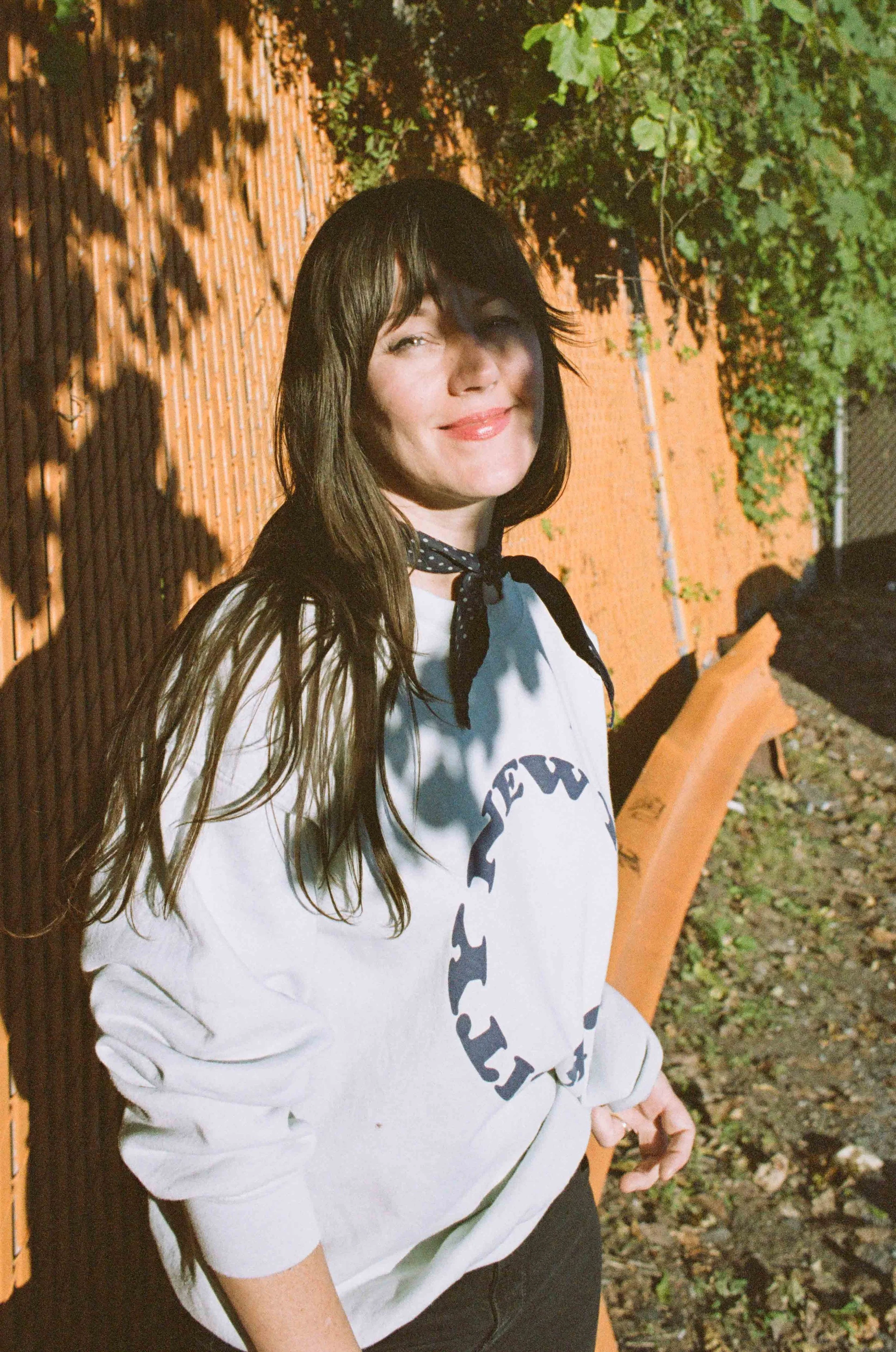 A woman with long brown hair and bangs, wearing a white sweatshirt with black text and a black and white polka dot scarf around her neck, standing outdoors at sunset against a brick fence with ivy and greenery. She is smiling softly and looking slightly to the side.