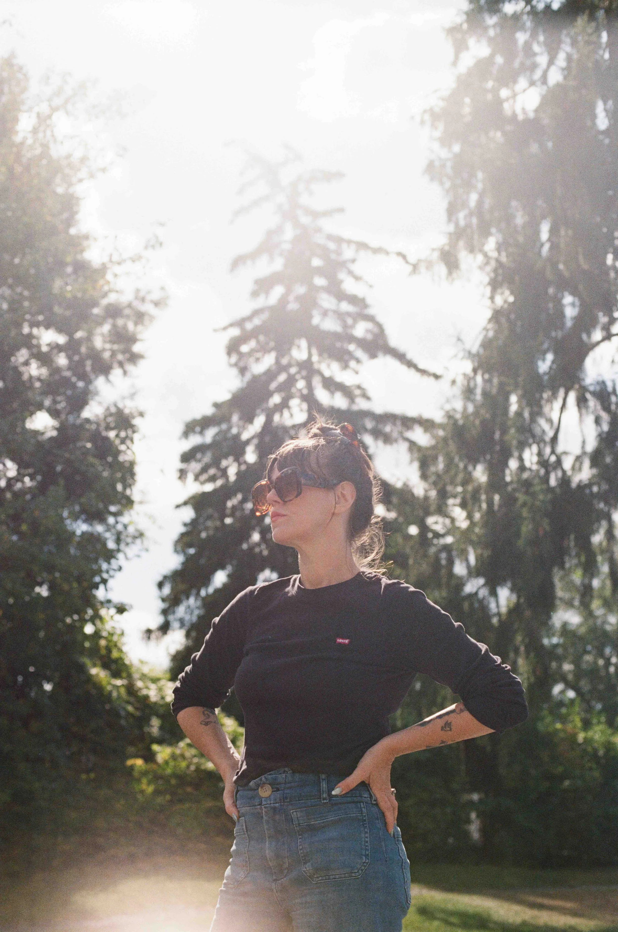 A woman stands outdoors with trees and sunlight behind her. She has brown hair and is wearing sunglasses, a black shirt, and jeans.