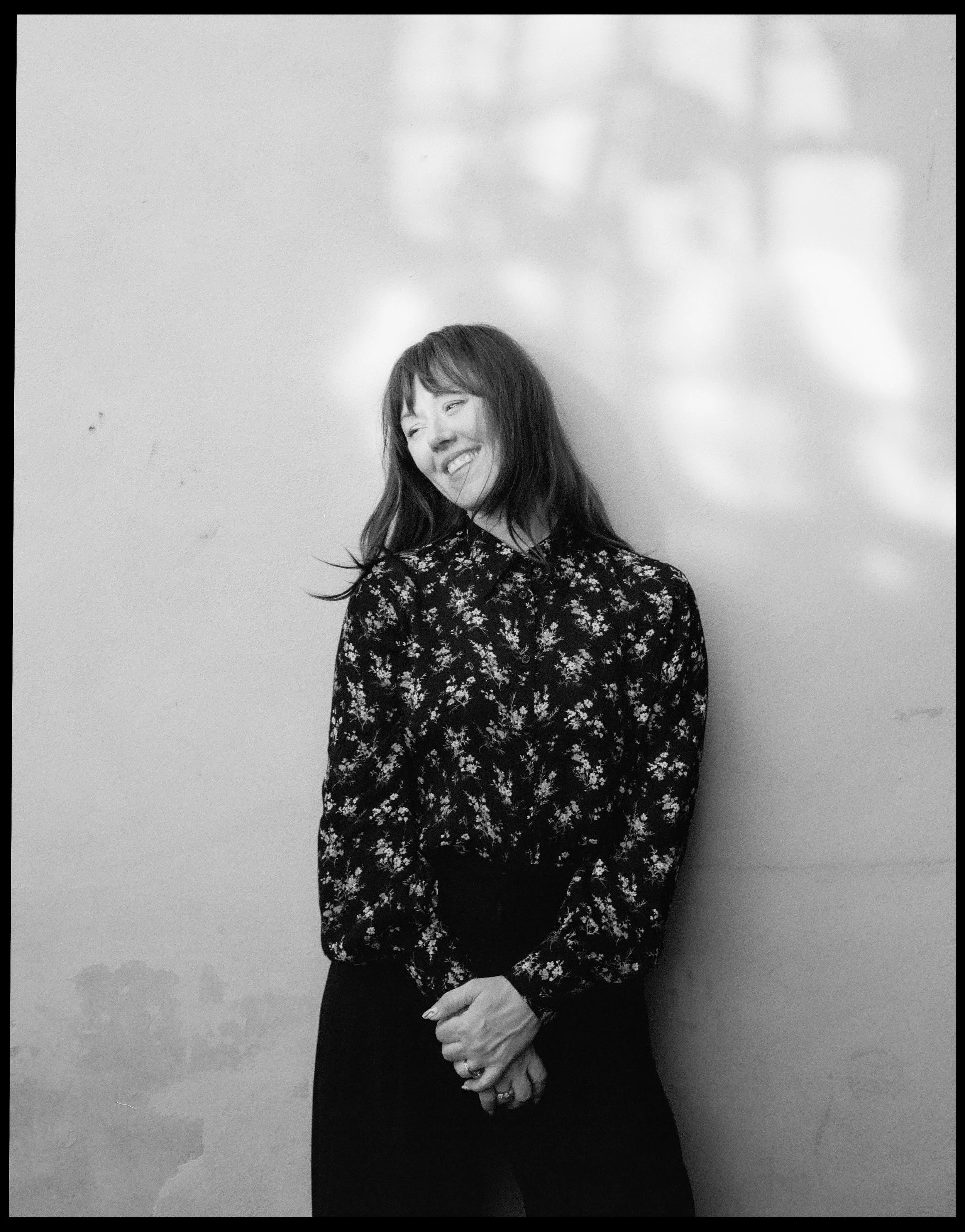 A woman with long hair, wearing a floral blouse, smiling and standing with her eyes closed against a plain wall with light reflections.