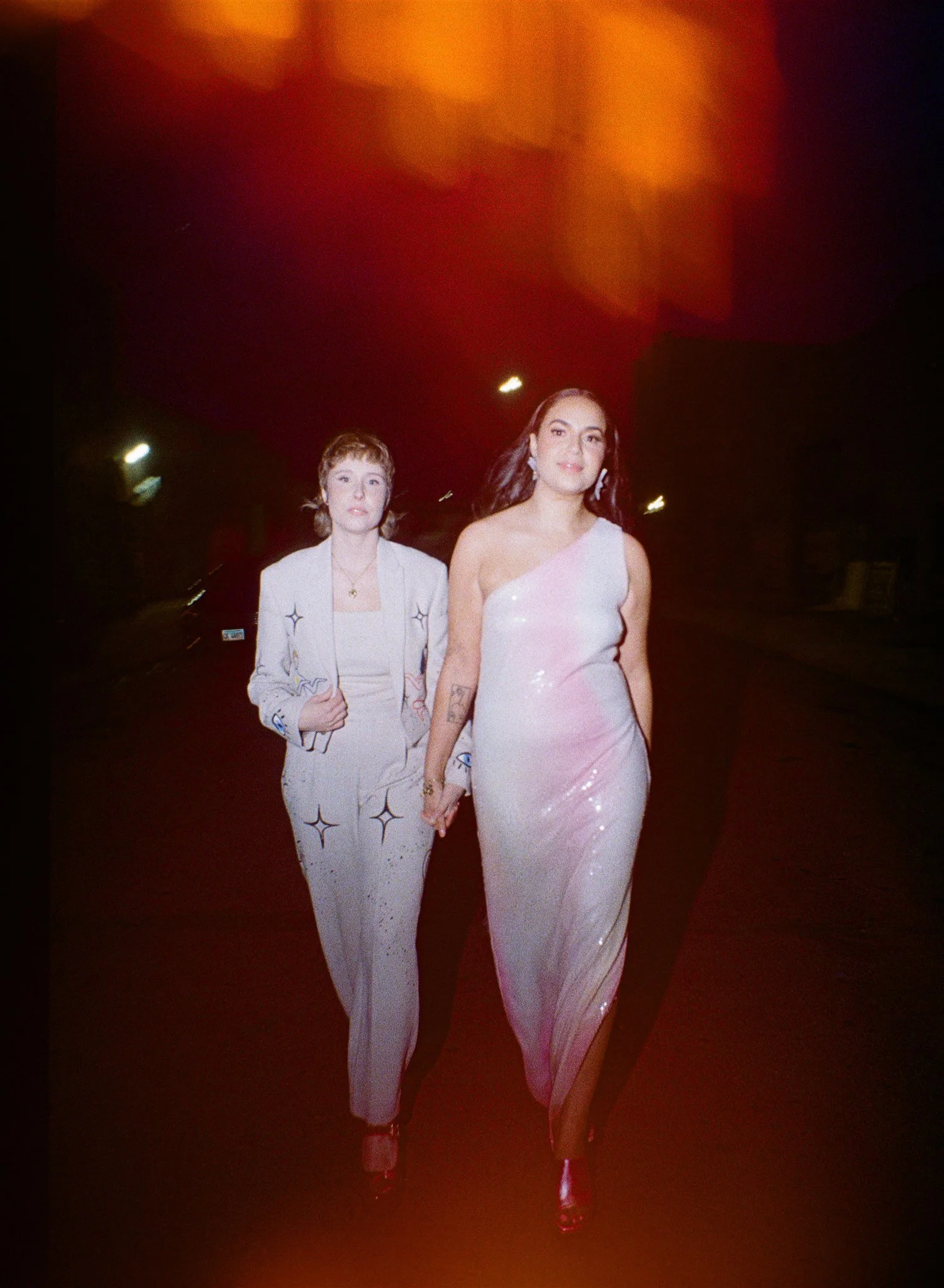 Two women walking hand in hand at night, one in a white suit with patterns and the other in a shiny, light pink dress.