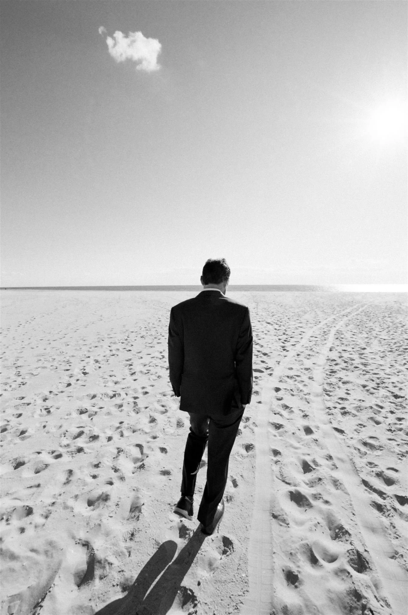 A man in a suit walking alone on a sandy beach with tire tracks and footprints, under a bright sky with a single cloud.