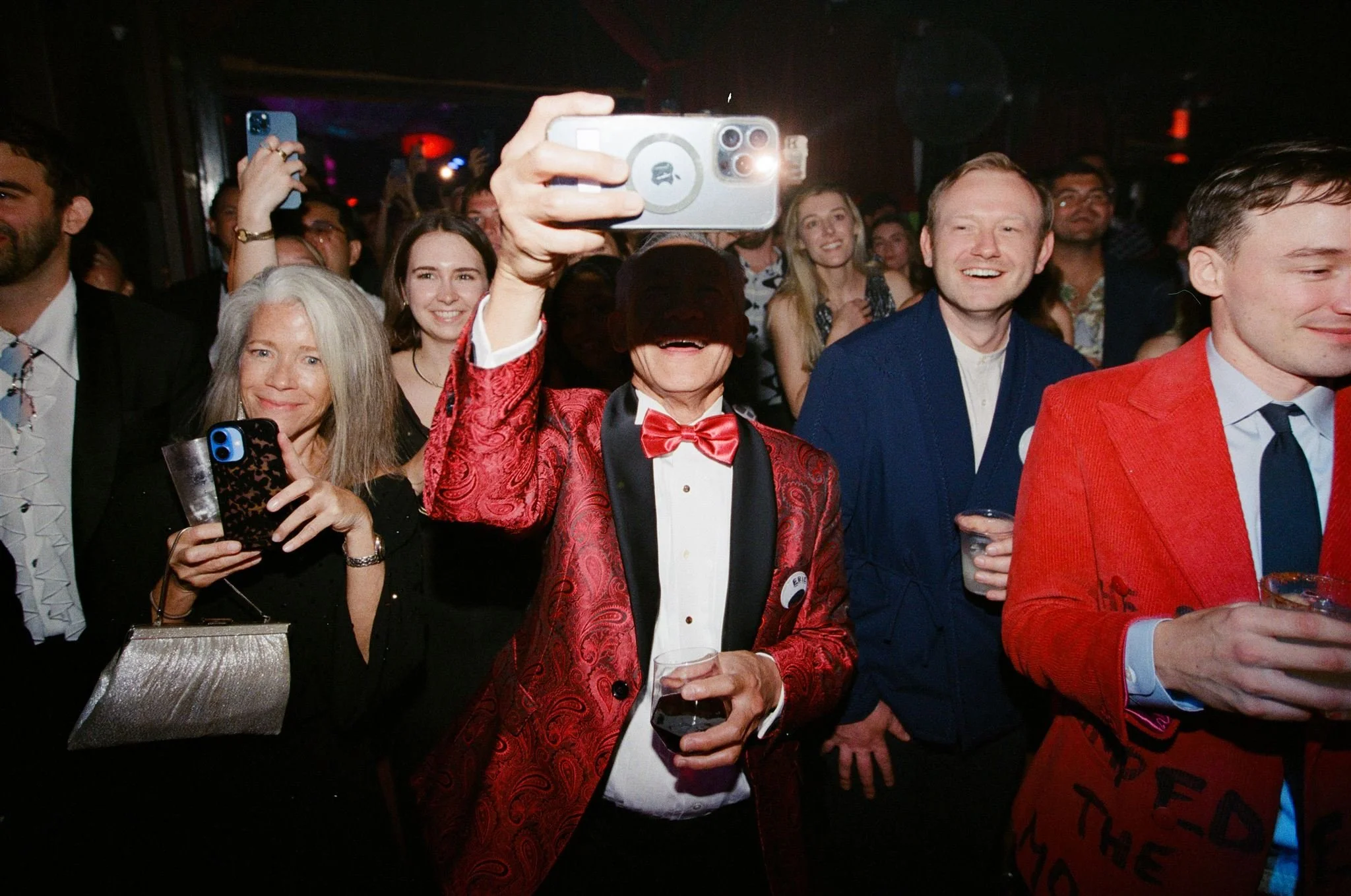 People at a lively party or celebration, with some taking photos and smiling, one person wearing a red blazer and bow tie, holding a drink.