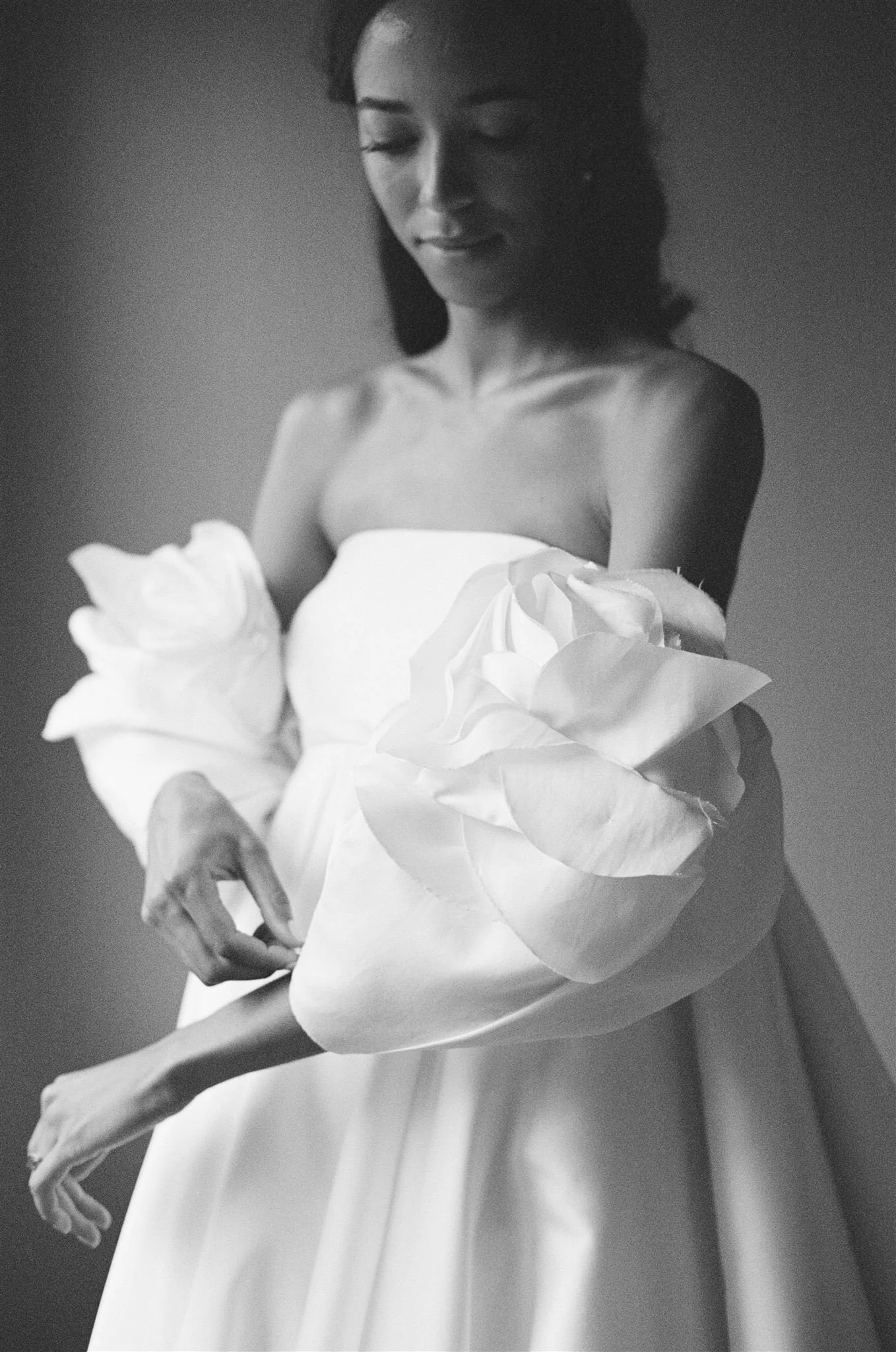 A black-and-white photo of a woman in a strapless dress holding a large flower with her arms extended.