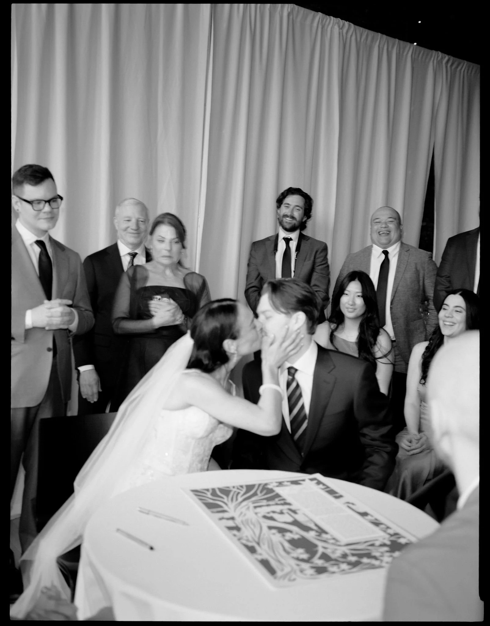A black-and-white photo of a wedding ceremony with the bride and groom kissing, surrounded by guests, some smiling and others watching. The bride is wearing a wedding dress and veil, while the groom is in a suit with a striped tie. The setting appear