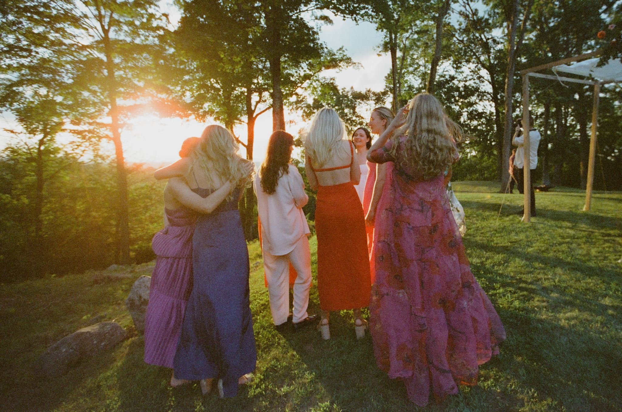 A group of women dressed in colorful dresses dancing together outdoors at sunset, with trees and a wooden structure in the background.