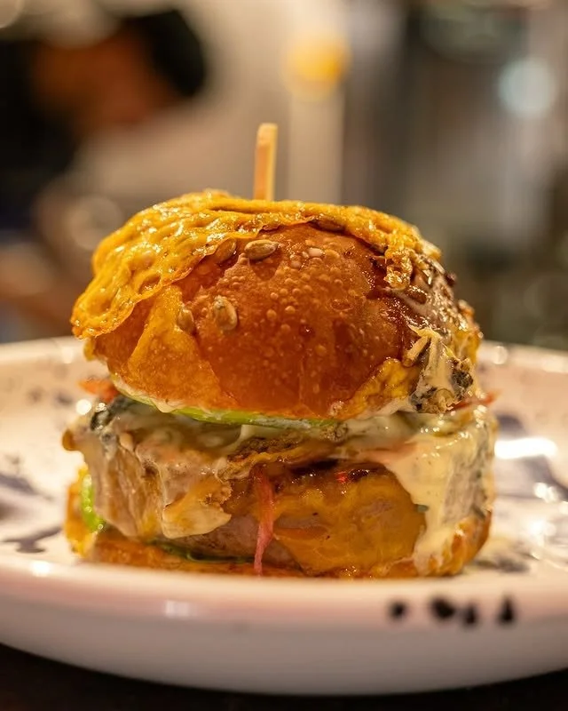 Un burger avec un petit pain, de la viande, du fromage, de la sauce, et une assiette blanche en arrière-plan.