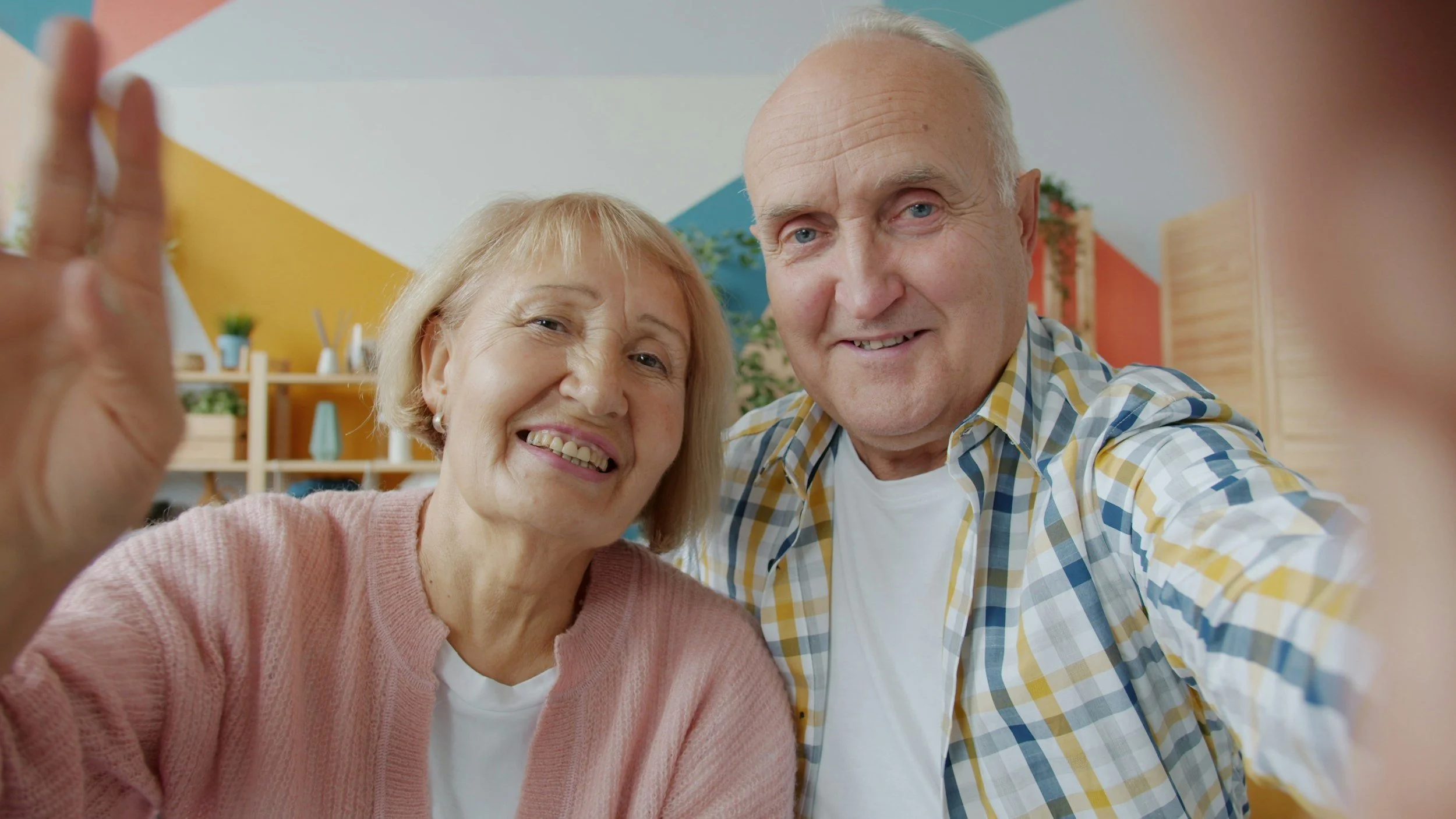 Two elderly people, a woman and a man, smiling and taking a selfie together in a brightly decorated room.
