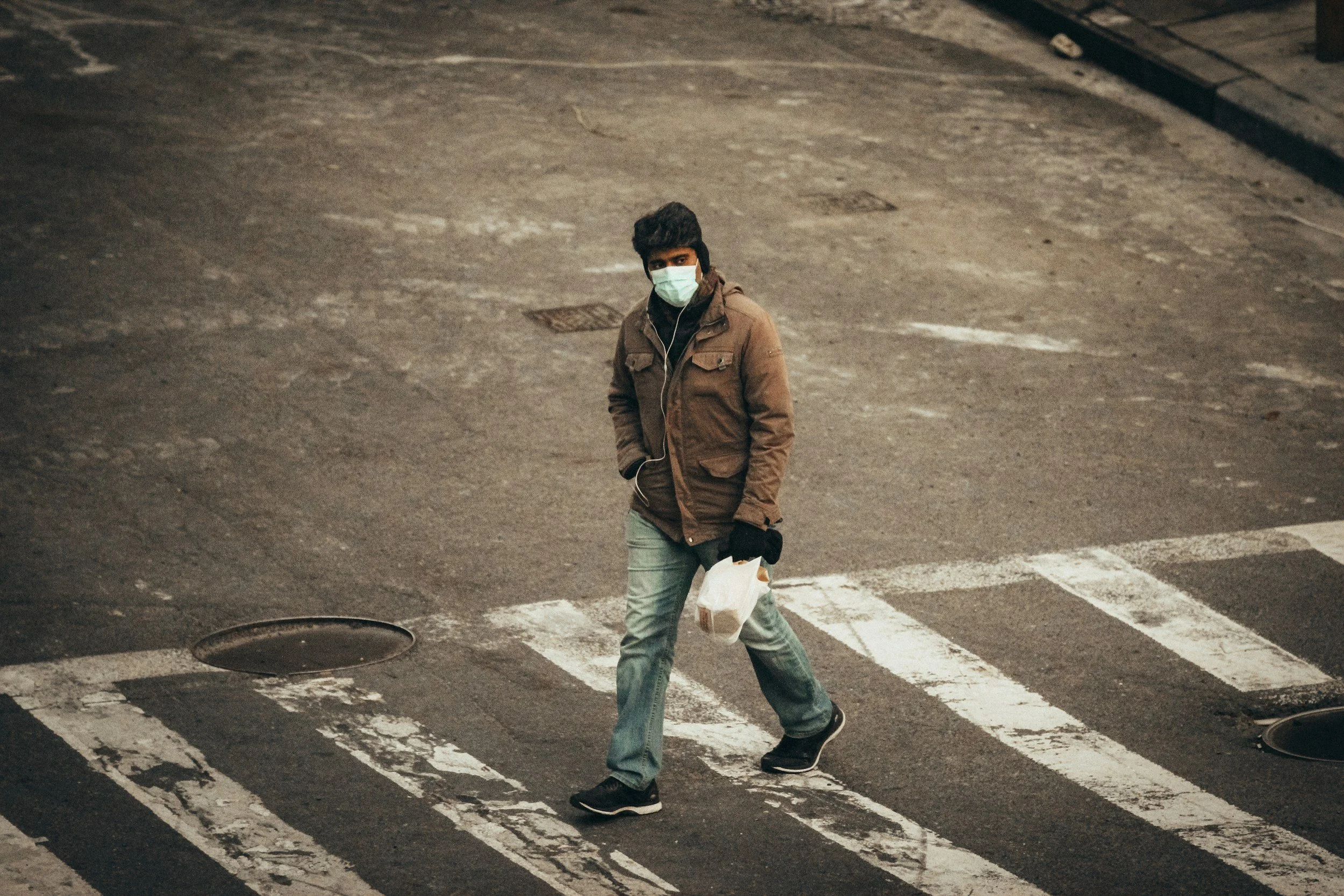 A man wearing a brown jacket, light jeans, black shoes, a face mask, and gloves crossing a city street at night, carrying a takeout food container.