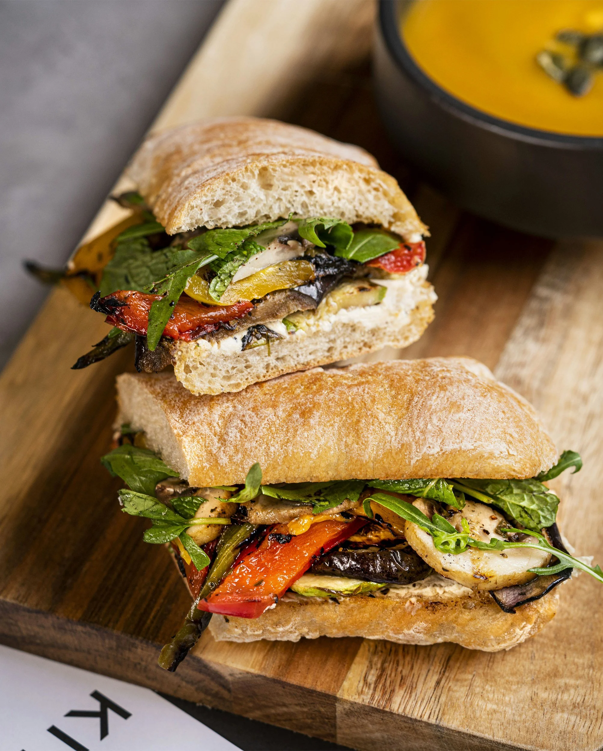 Two sandwiches with roasted vegetables and greens on crusty bread on a wooden board, with a bowl of soup in the background.