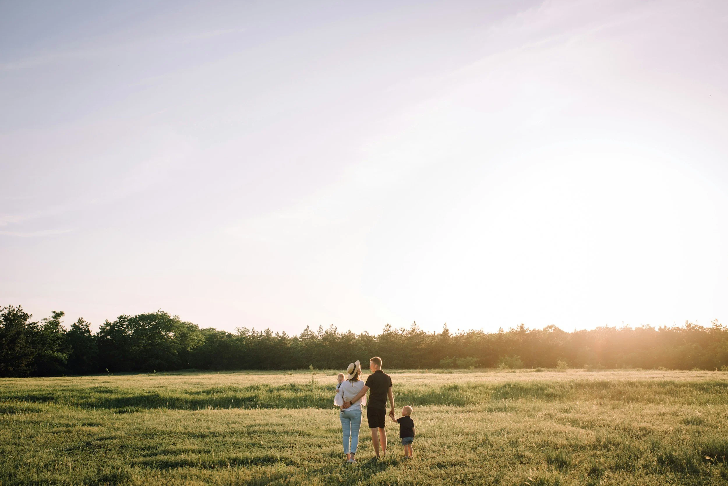 A family of four walking hand-in-hand in a field during sunset, with a line of trees in the background.