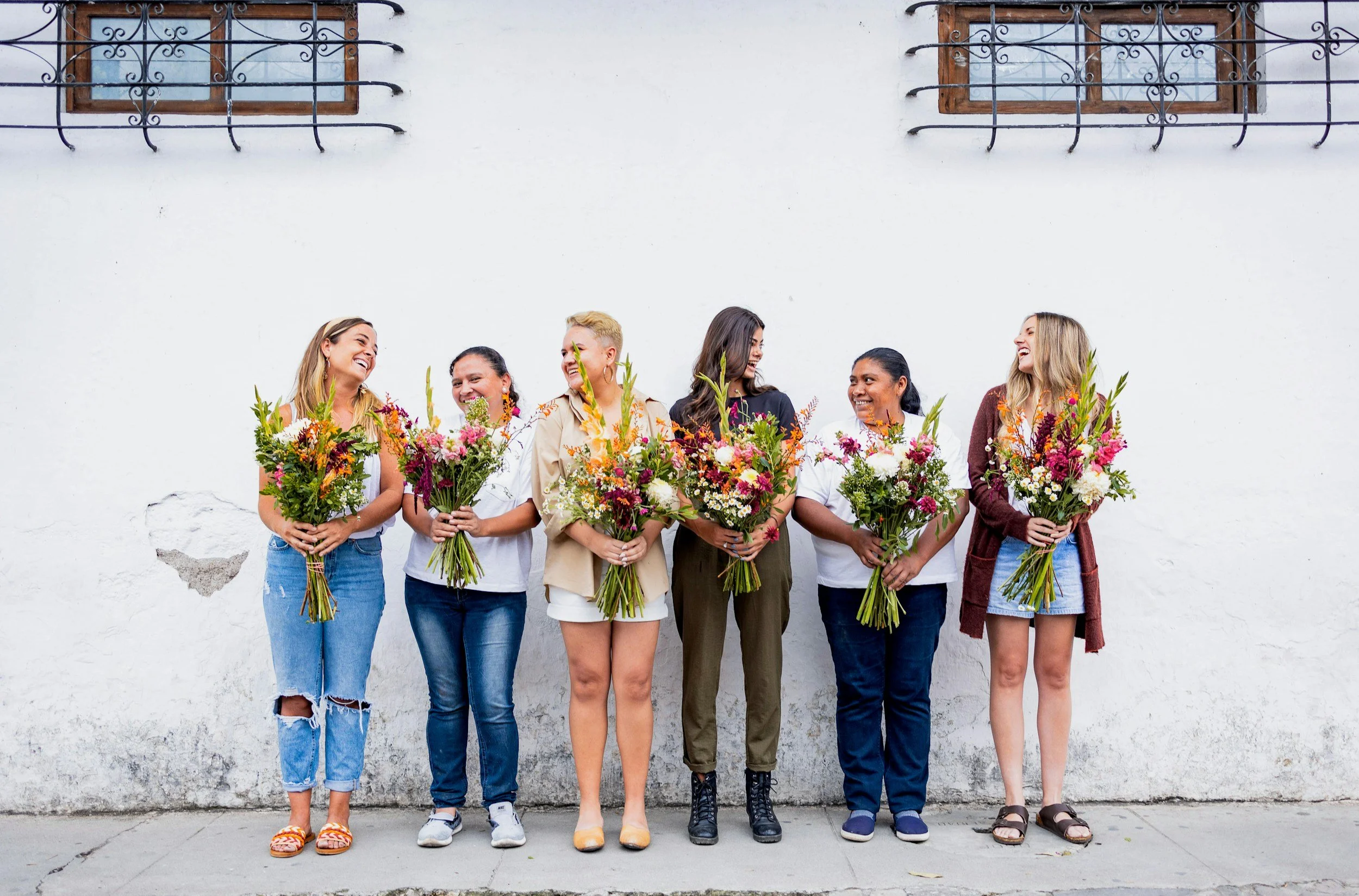 A group of six smiling women standing against a white wall, each holding a bouquet of colorful flowers.
