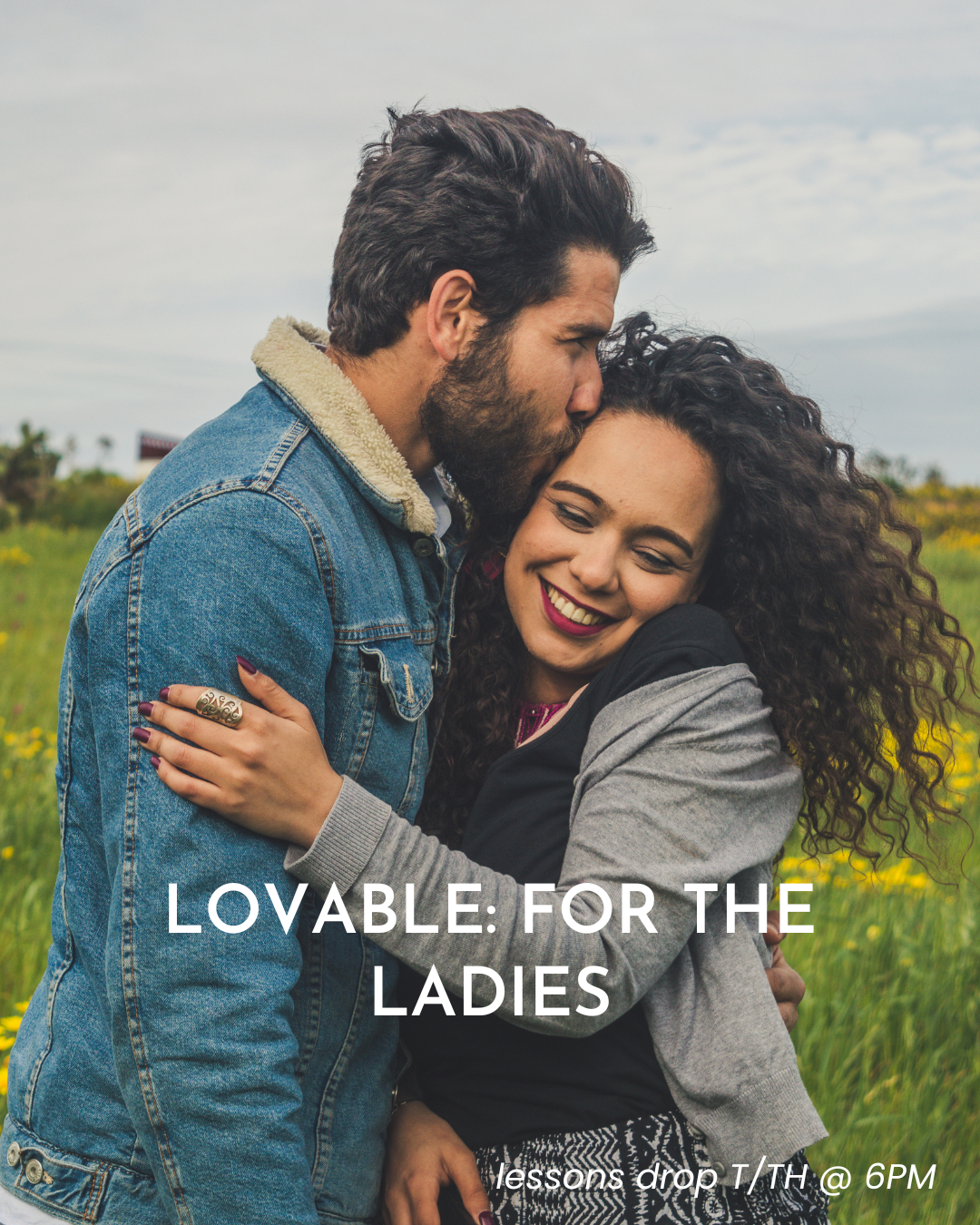 A man and woman hugging outdoors in a field with yellow flowers. The man is kissing the woman on the forehead, and she is smiling with eyes closed. The woman has curly hair and wears a gray and black jacket. The man has dark hair, a beard, and is wearing a denim jacket with a shearling collar.