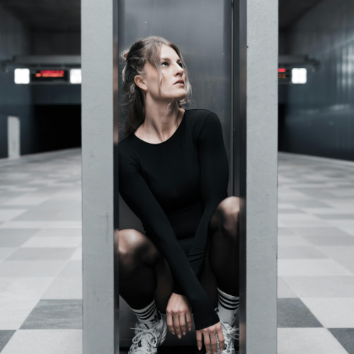 A woman with long brown hair in a black long-sleeve top and black shorts, sitting on the floor with knees up, in a small enclosed space or telephone booth, looking to the side in an underground or subway station.