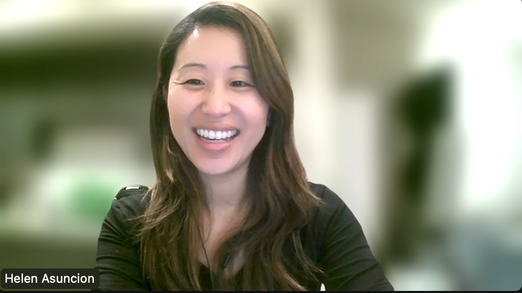 A woman with long brown hair smiling during a video call, wearing a black top, in front of a blurred background.