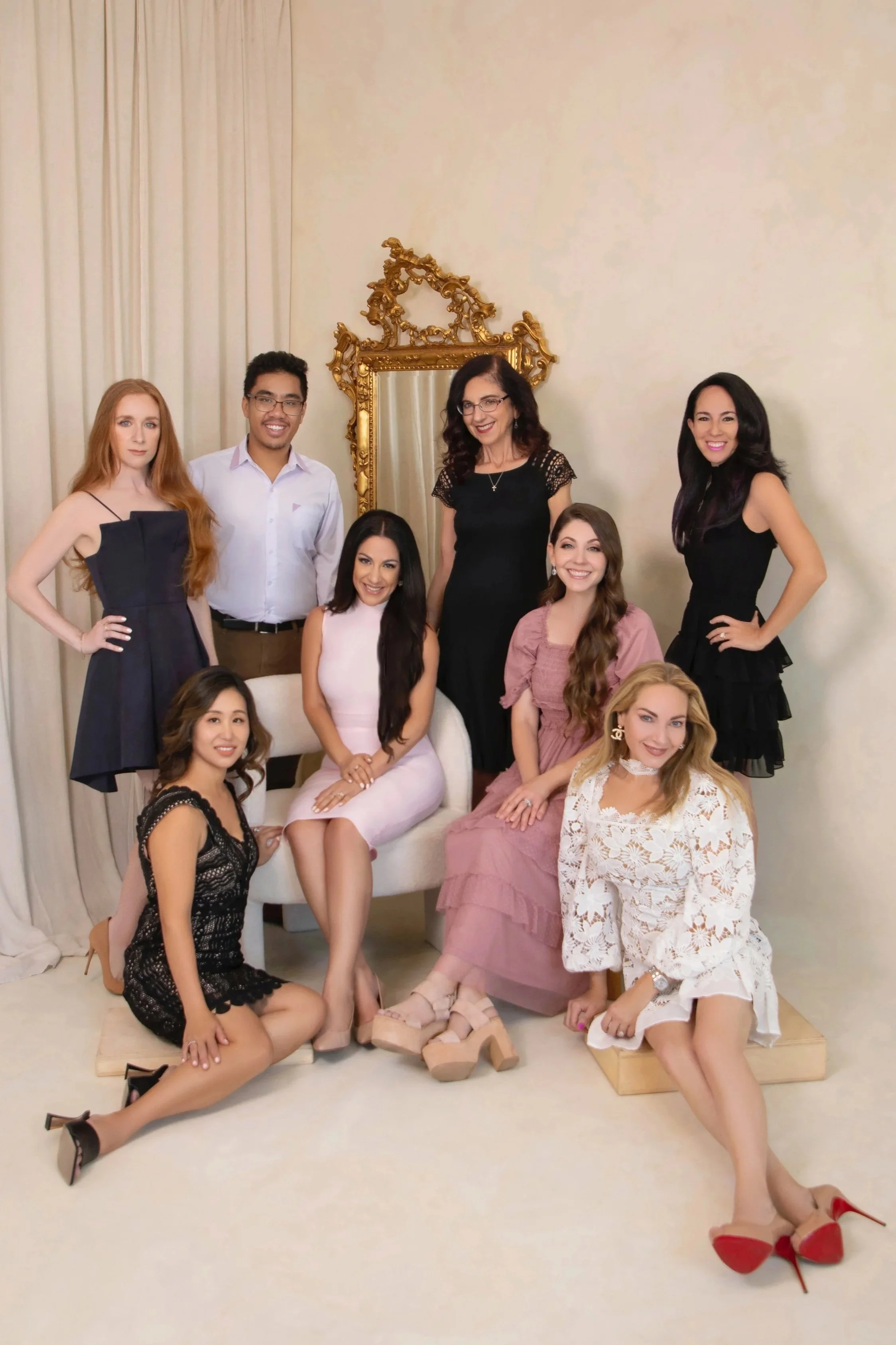 Group of nine people, seven women and two men, dressed elegantly in a room with beige walls and an ornate gold-framed mirror, posing for a photo.