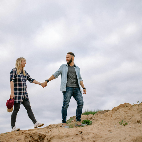 A young couple holding hands and smiling at each other outdoors on a cloudy day, standing on a sandy hill.