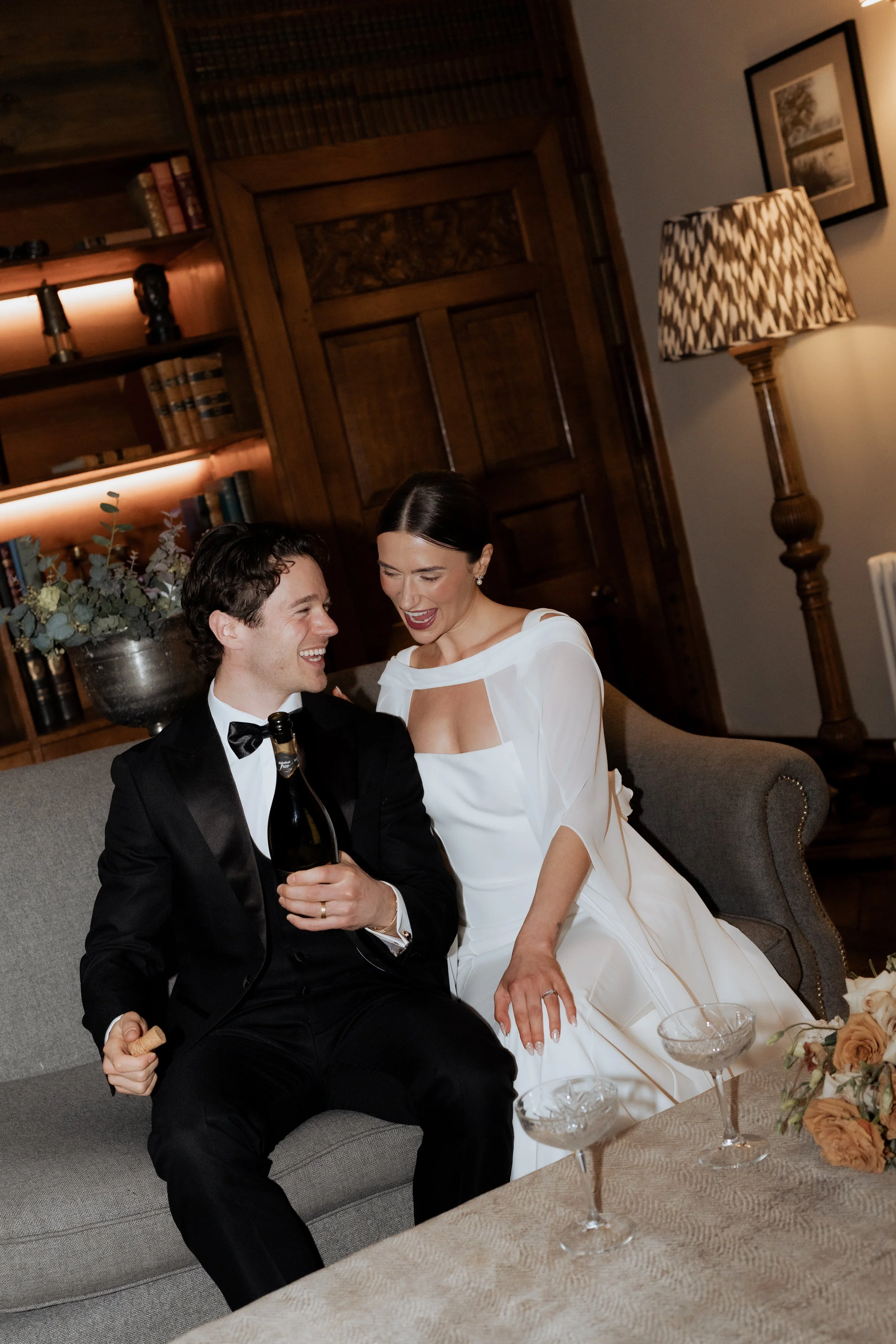 Bride and groom relaxing together on a sofa at Soughton Hall, sharing champagne during an editorial wedding moment in soft, ambient light.