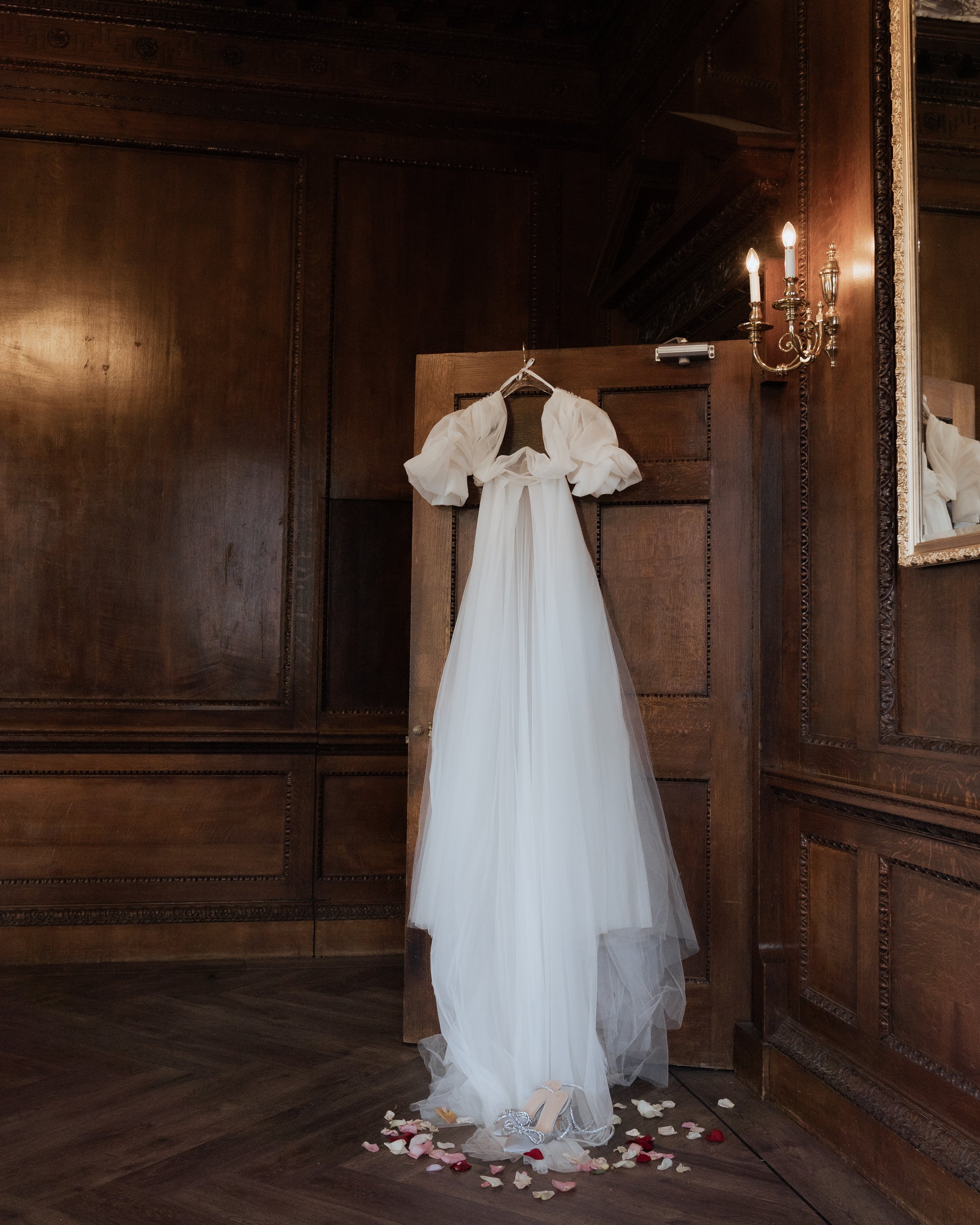 Editorial wedding dress hanging on a wooden door inside a dark, elegant Staffordshire wedding venue, styled with scattered rose petals and soft romantic bridal details.