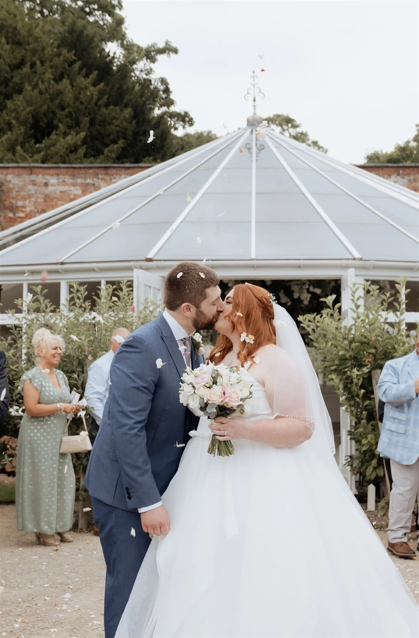 Bride and groom sharing a confetti kiss outside the glasshouse at Combermere Abbey during an elegant Cheshire wedding.