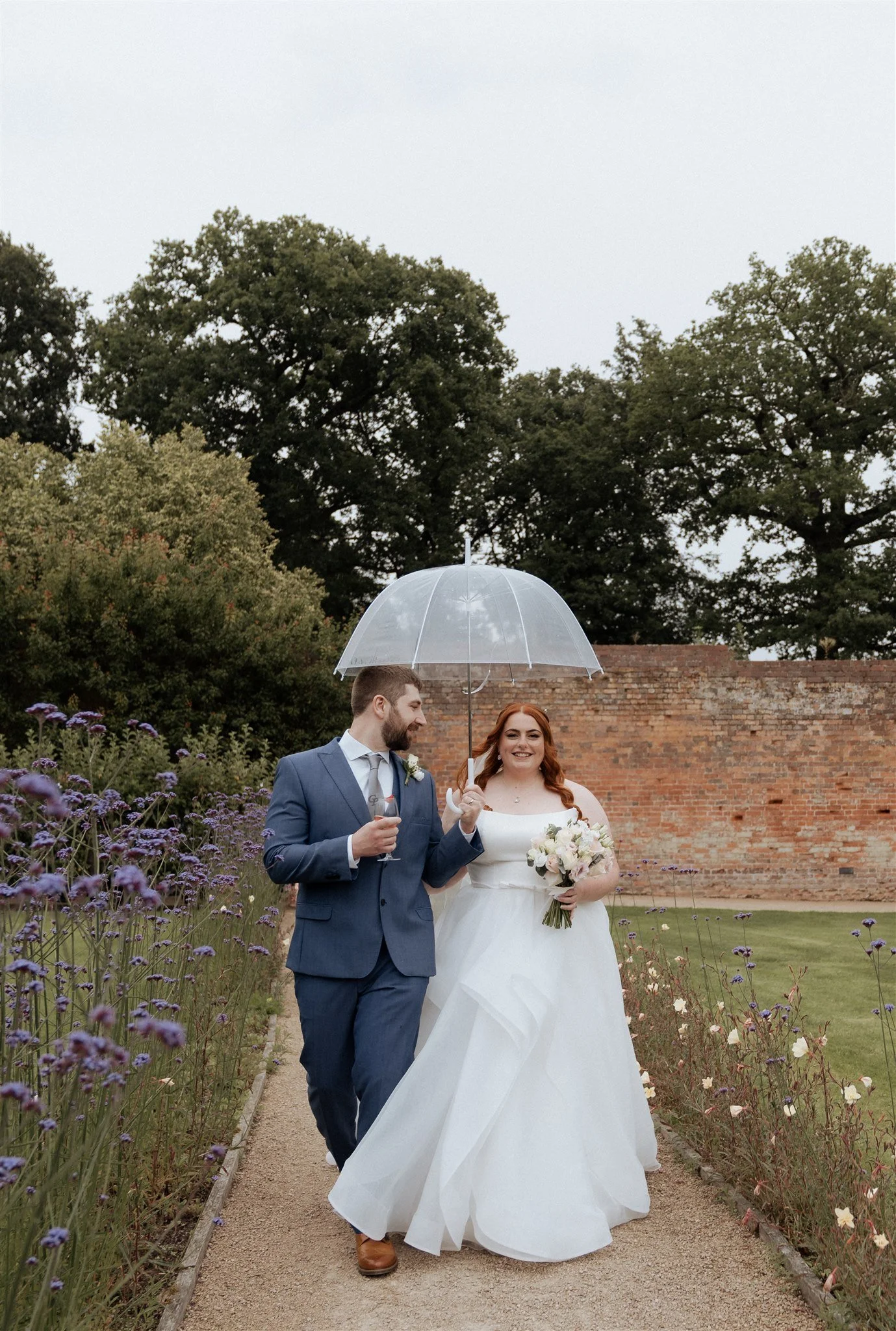 Bride and groom walking through the gardens at Combermere Abbey with a clear umbrella during an elegant countryside wedding