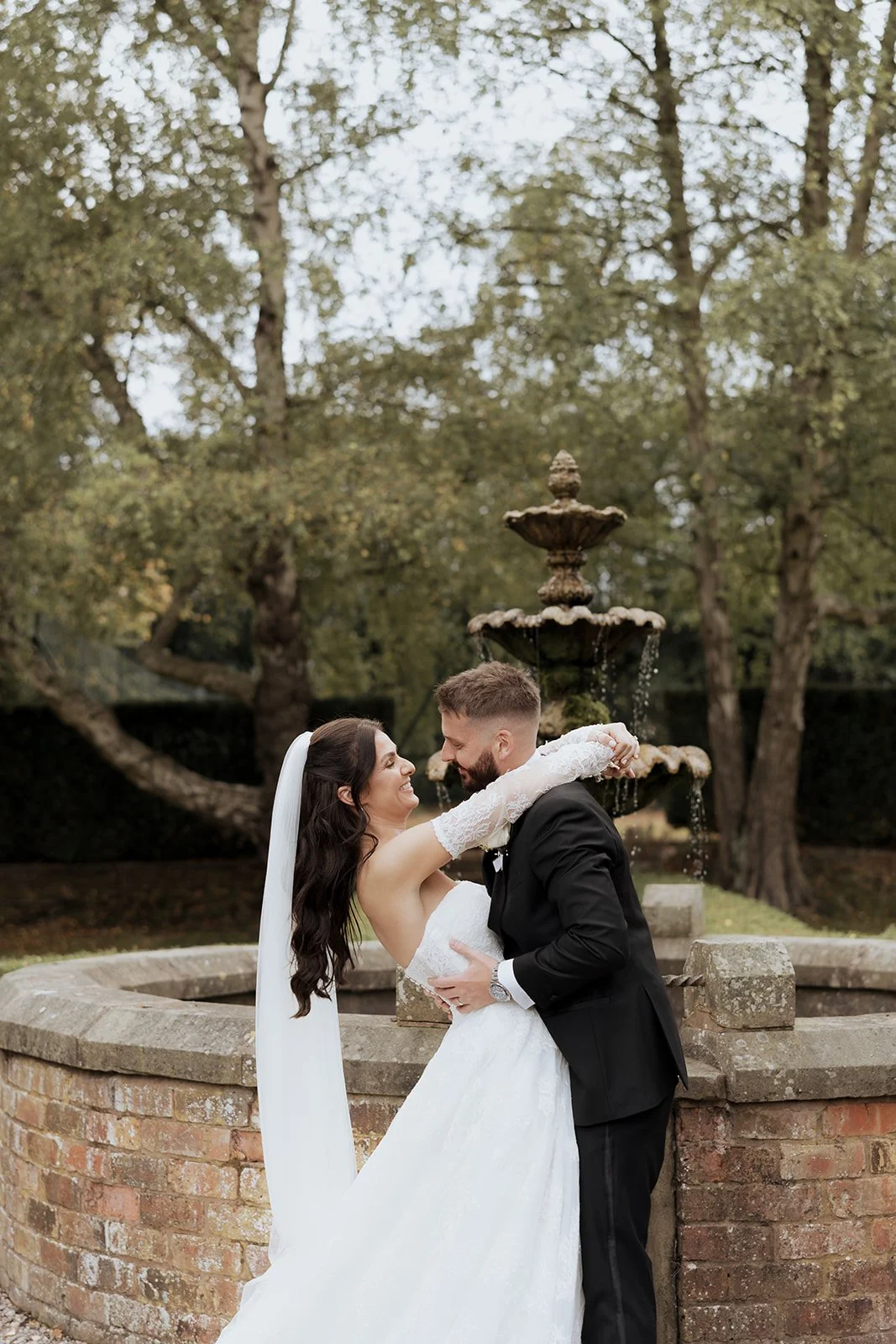 Bride and groom embracing in front of the garden fountain at Soughton Hall, surrounded by trees with soft natural light and timeless editorial wedding styling.