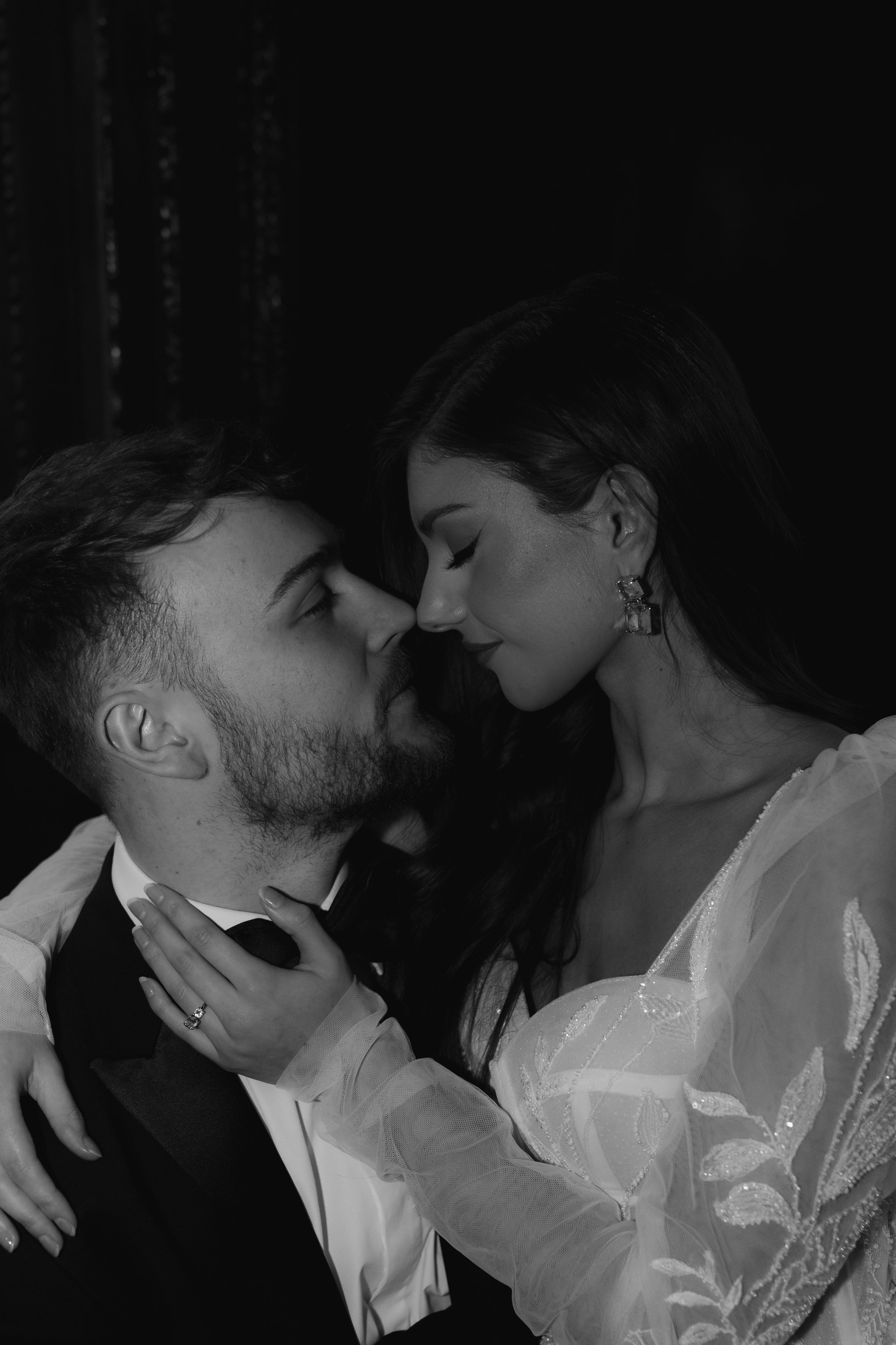 Black and white editorial wedding portrait of a bride and groom sharing an intimate moment, captured in soft dramatic light with the bride’s embroidered gown and engagement ring visible.