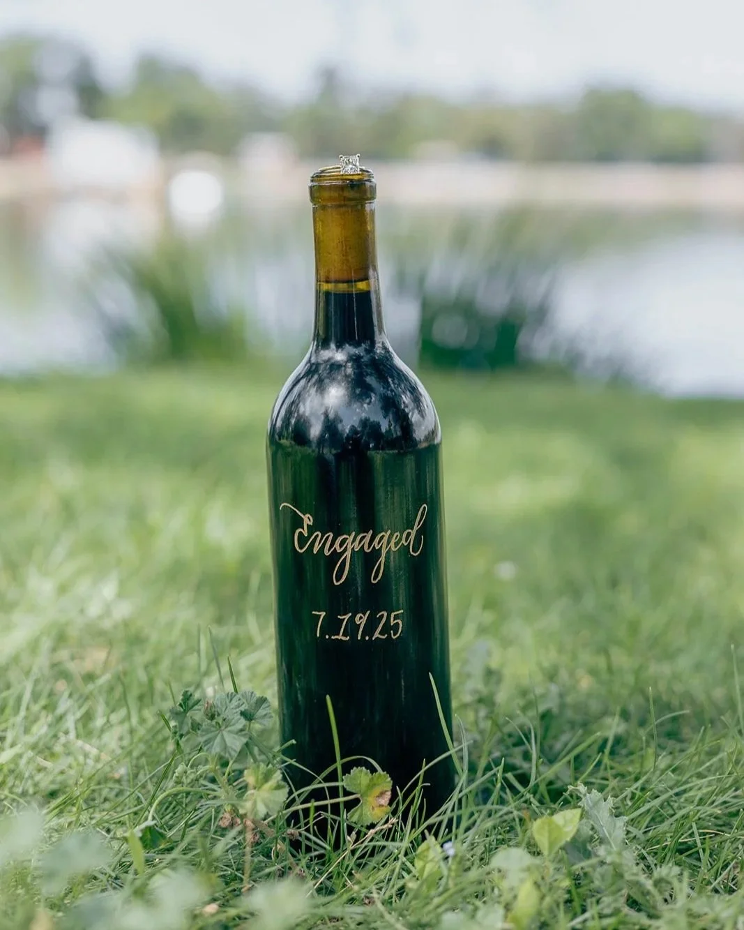 A green glass bottle standing upright outdoors on grass, engraved with the words 'Engaged' and the date '7.19.25', with a wedding ring on top of the bottle's opening.