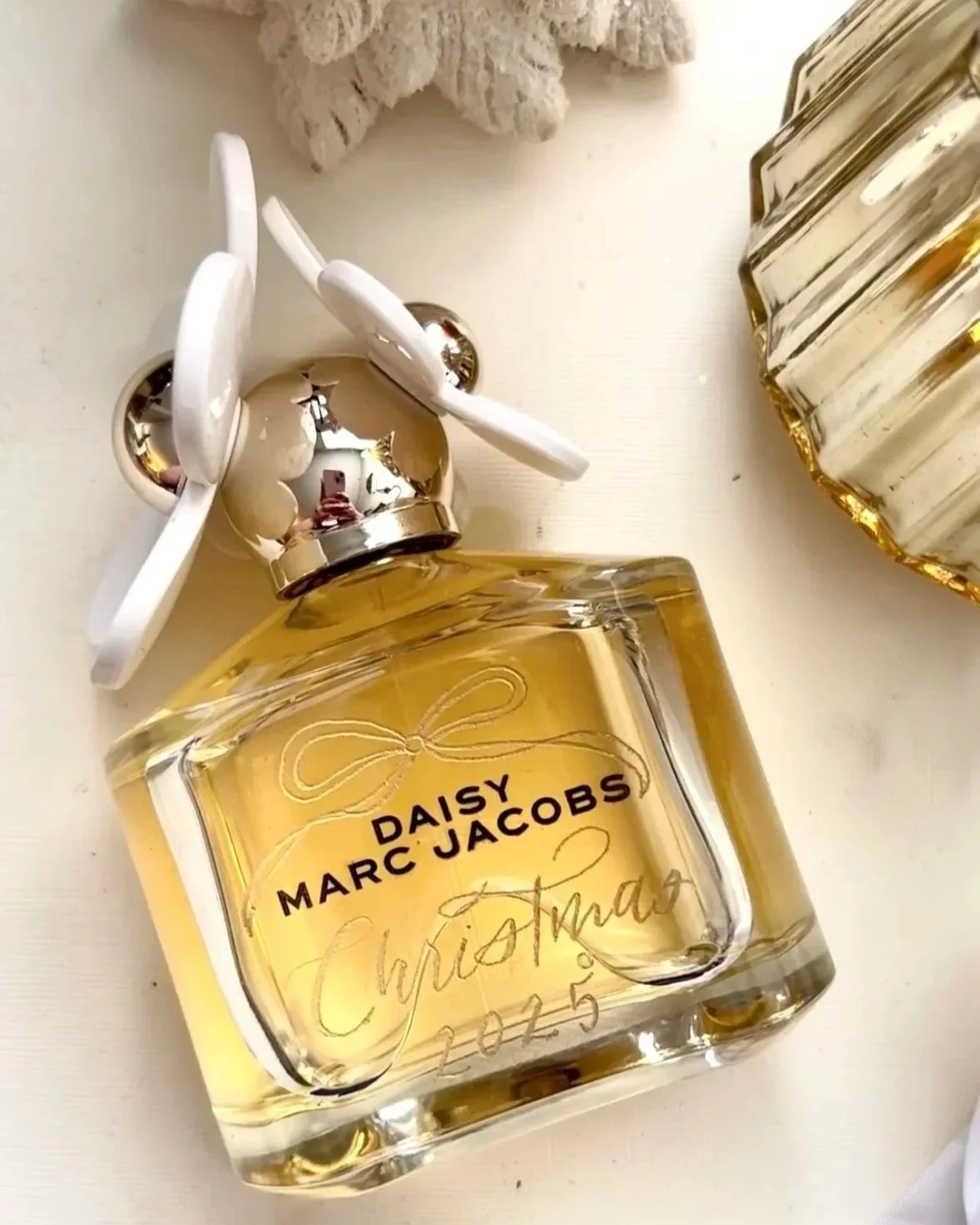 On-site personalization in Denver, CO on Daisy Marc Jacobs perfume bottle with a silver bow on its cap, placed on a white surface, with another decorative glass item in the background.