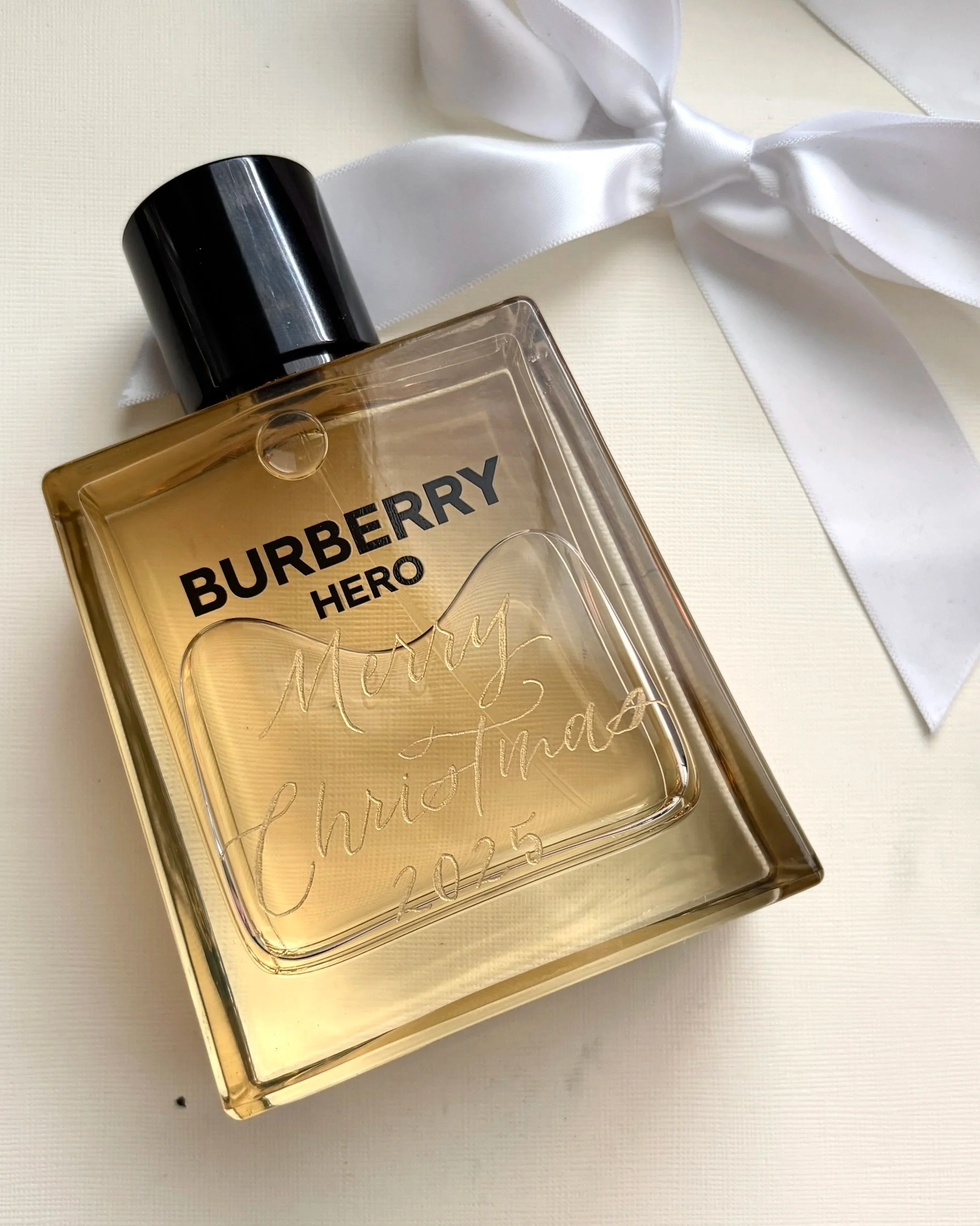 Live, onsite calligraphy in Denver, CO on A Burberry perfume bottle with etched inscription, resting on a white surface next to a white ribbon tied in a bow.